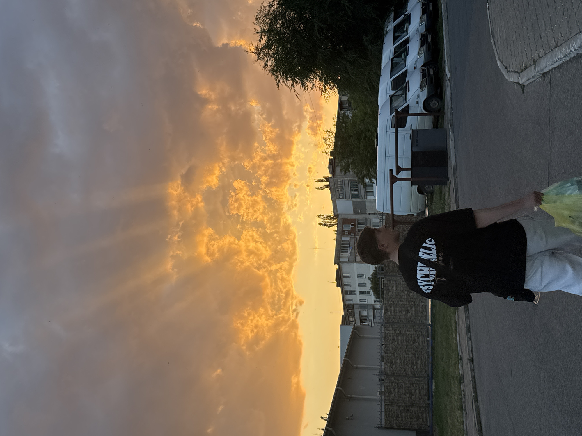 A young man in a black t-shirt and white pants walks away from the camera on a paved street in Tiraspol, Moldova. He carries a yellow plastic bag in his right hand. The sky is filled with dramatic orange and grey clouds illuminated by the setting sun. In the background, a row of white vans is parked in front of apartment buildings and a tree. A fence borders a grassy area to the left of the street.