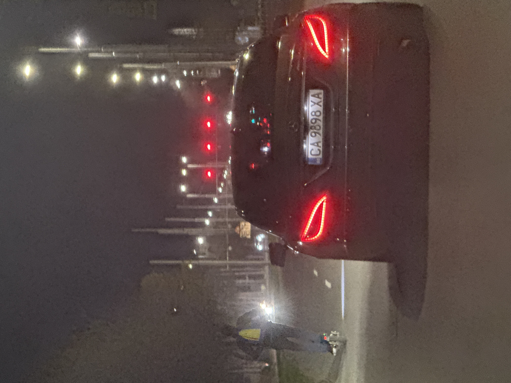 The image captures a nighttime scene in Plovdiv, Bulgaria. A dark-colored car with illuminated tail lights is prominently featured, displaying the license plate "CA 9898 XA". A person wearing a reflective safety vest and light-colored sneakers is standing beside the road. The backdrop includes street lights lining the road and visible red lights, possibly traffic signals. The surroundings suggest an urban environment.