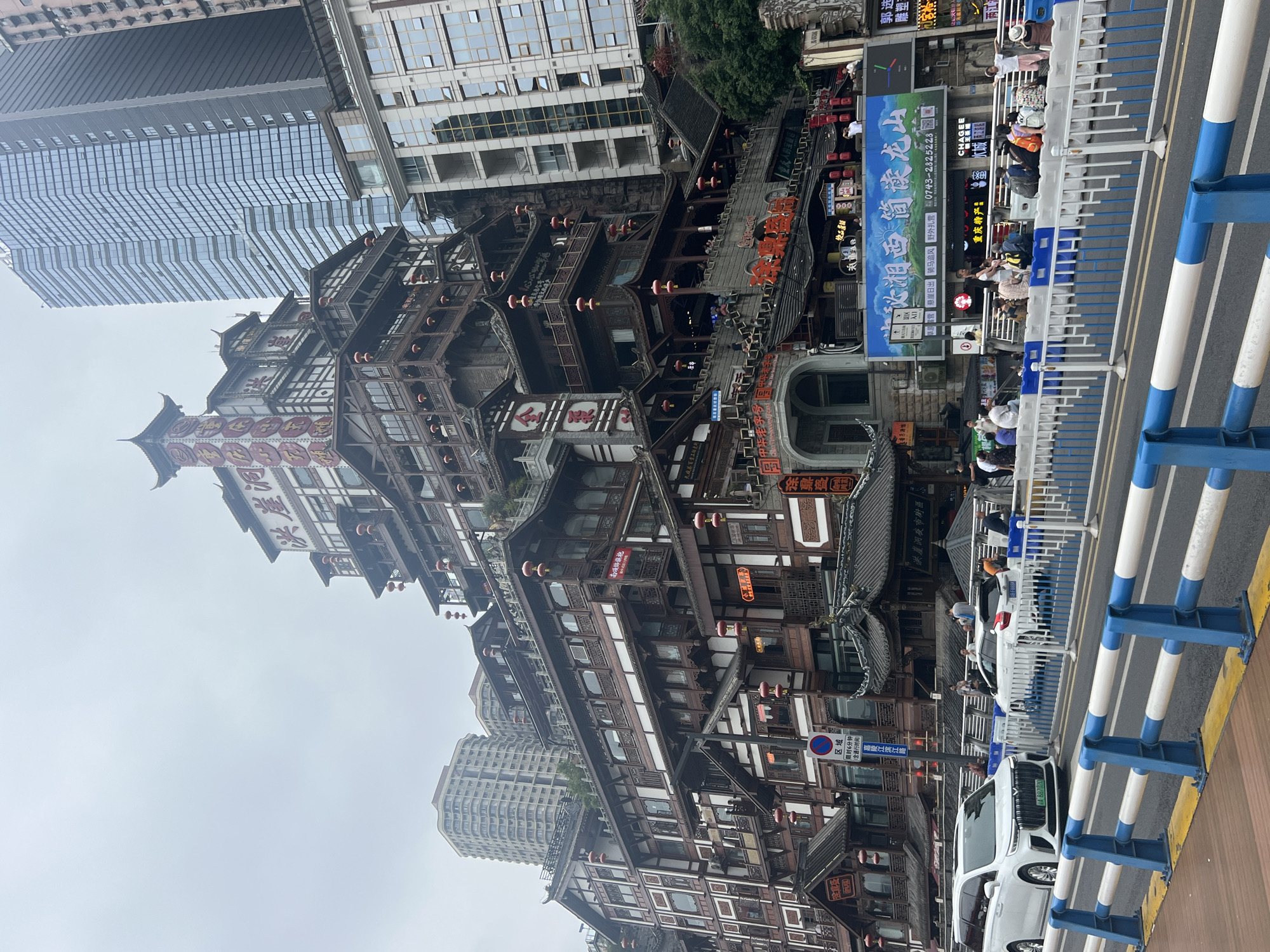 The image shows a multi-story building with traditional Chinese architecture, possibly a hotel or a commercial complex, in the Yuzhong District of China. The building has a tiered structure with numerous balconies, wooden accents, and red lanterns. It is situated on a hillside and appears to be a prominent landmark. In the foreground, a road with a white and blue striped barrier runs across the image. Several vehicles, including a white minivan and a white car, are parked or driving on the road. Pedestrians are visible on the sidewalk and on the building's balconies, some appearing to be tourists. The sky is overcast, suggesting a cloudy day. Modern skyscrapers are visible in the background, contrasting with the traditional building. A large billboard with Chinese text is displayed on the side of the building.