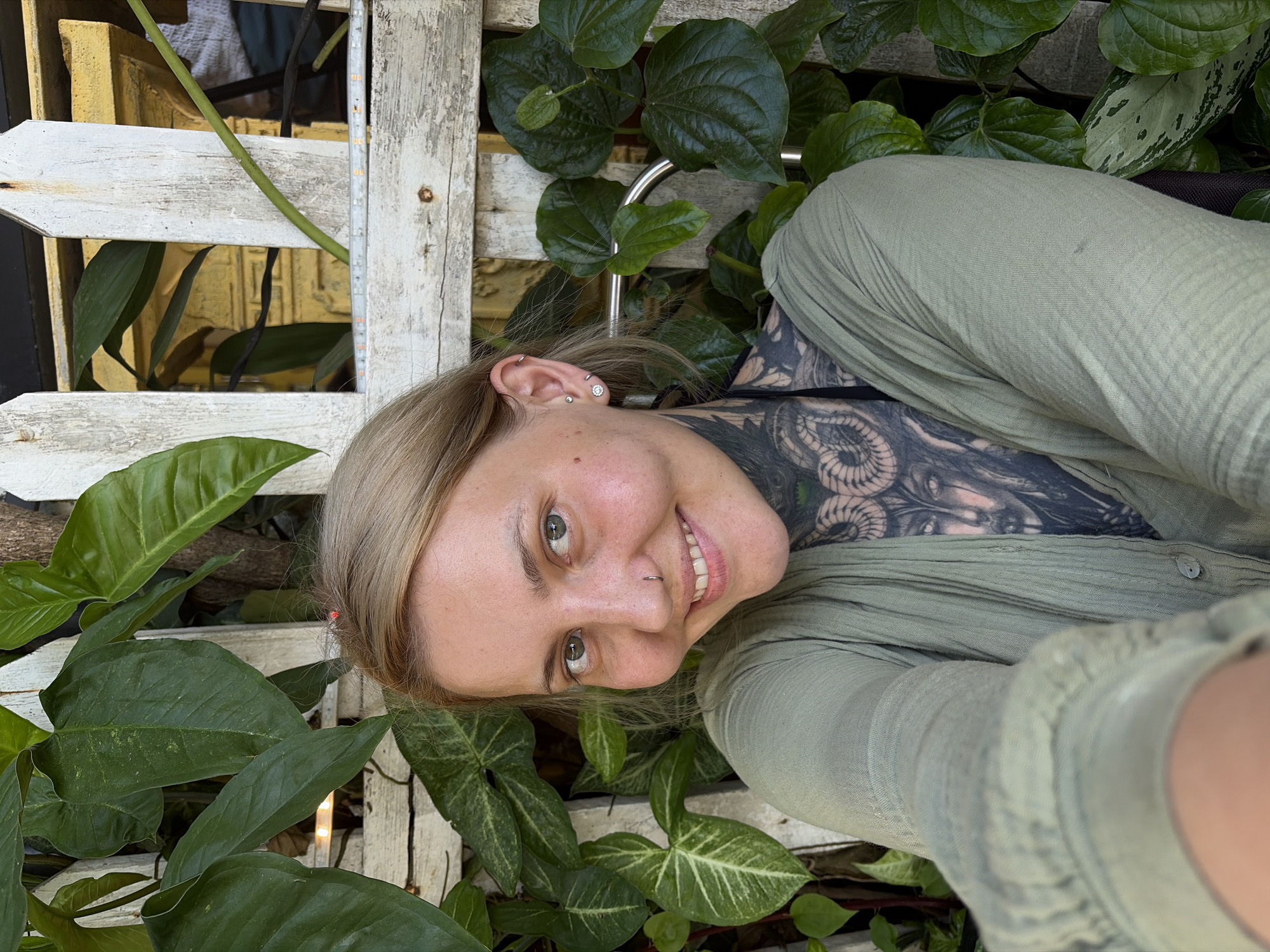 A blonde woman with light-colored eyes is captured in a close-up, eye-level shot, smiling warmly and looking directly at the camera. Her head is tilted, giving the impression of a candid moment or a selfie. She has a small hoop piercing in her nose and multiple stud and hoop piercings in her left ear. Extensive black and grey tattoos adorn her neck, chest, and shoulder, featuring intricate designs that include swirling patterns and what appears to be a stylized face or skull. She is wearing a light green, possibly linen, collared shirt.
The background is dominated by lush green foliage of various plants, characteristic of a tropical environment like Hội An Ward, Vietnam. Some leaves are plain dark green, while others display lighter patterns. A weathered white wooden fence or trellis structure is prominent, running horizontally and vertically through the scene. A thin strip of illuminated LED lights is visible vertically on one of the fence posts. Hints of yellow structures can be seen further in the background, behind the fence and plants. The natural lighting suggests it is daytime. The woman's expression conveys happiness and comfort in her surroundings. There is no visible text in the image.