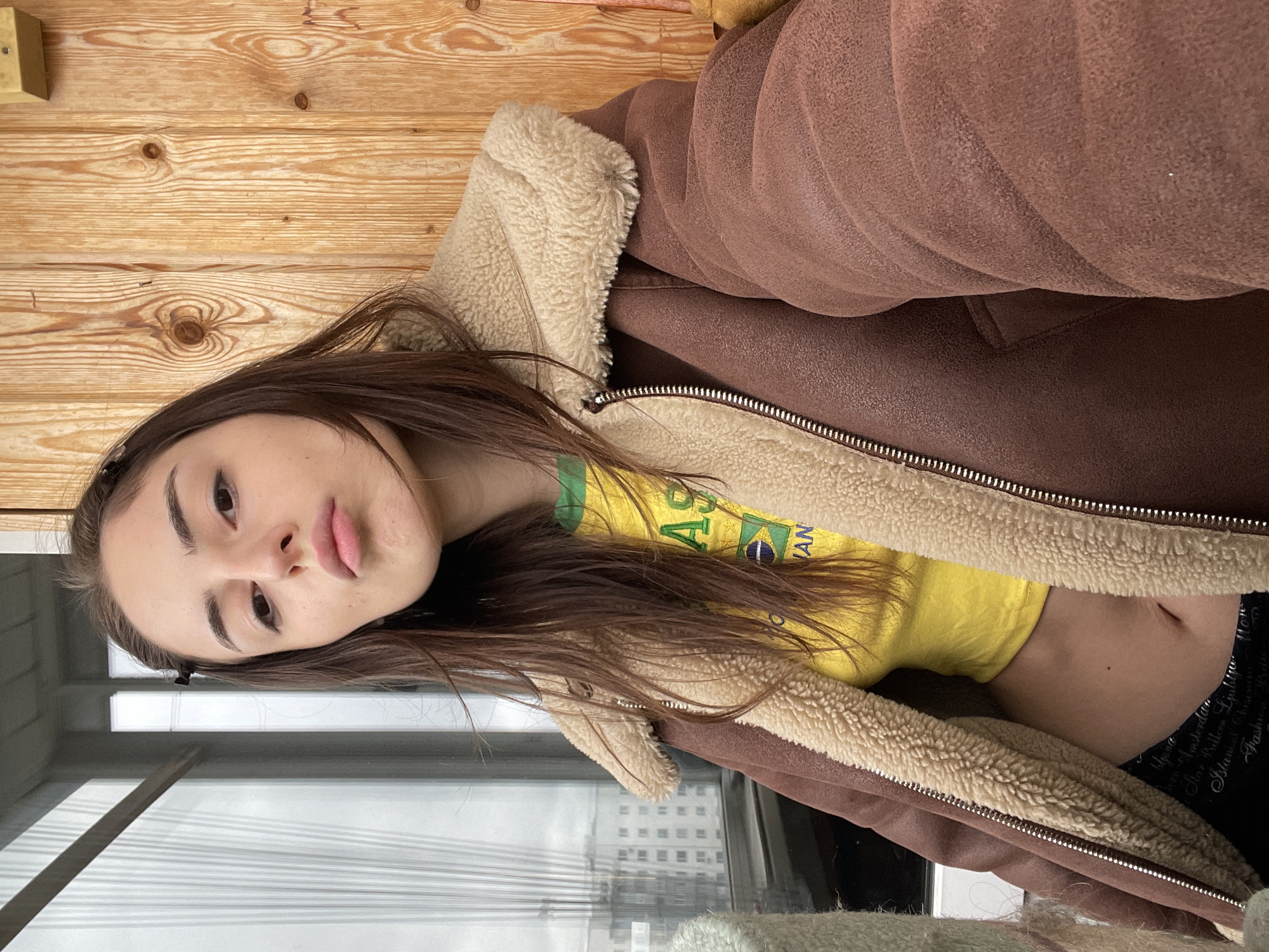 A young woman with long, dark hair poses for a photograph indoors, with her head tilted to the side. She is wearing a yellow t-shirt featuring the Brazilian flag and partial text that includes "BRASIL". Over this, she wears a brown, suede-like jacket with a cream-colored shearling lining, partially unzipped to reveal her midriff. Her expression is neutral, with slightly pouted lips.
The background behind her head is a wooden plank wall, showing natural wood grain. To her right, the folds and texture of her jacket are visible. To her left and below, a large window offers a view of a city skyline, including distant buildings and what appears to be the structural cables of a bridge or similar large construction, indicating an urban setting. The lighting suggests it is daytime. Her dark bottom wear has faint white text, with visible words like "Amsterdam," "Star Killer," and "Fashion." The overall scene suggests a casual moment in an apartment or room in Moscow, Russia.