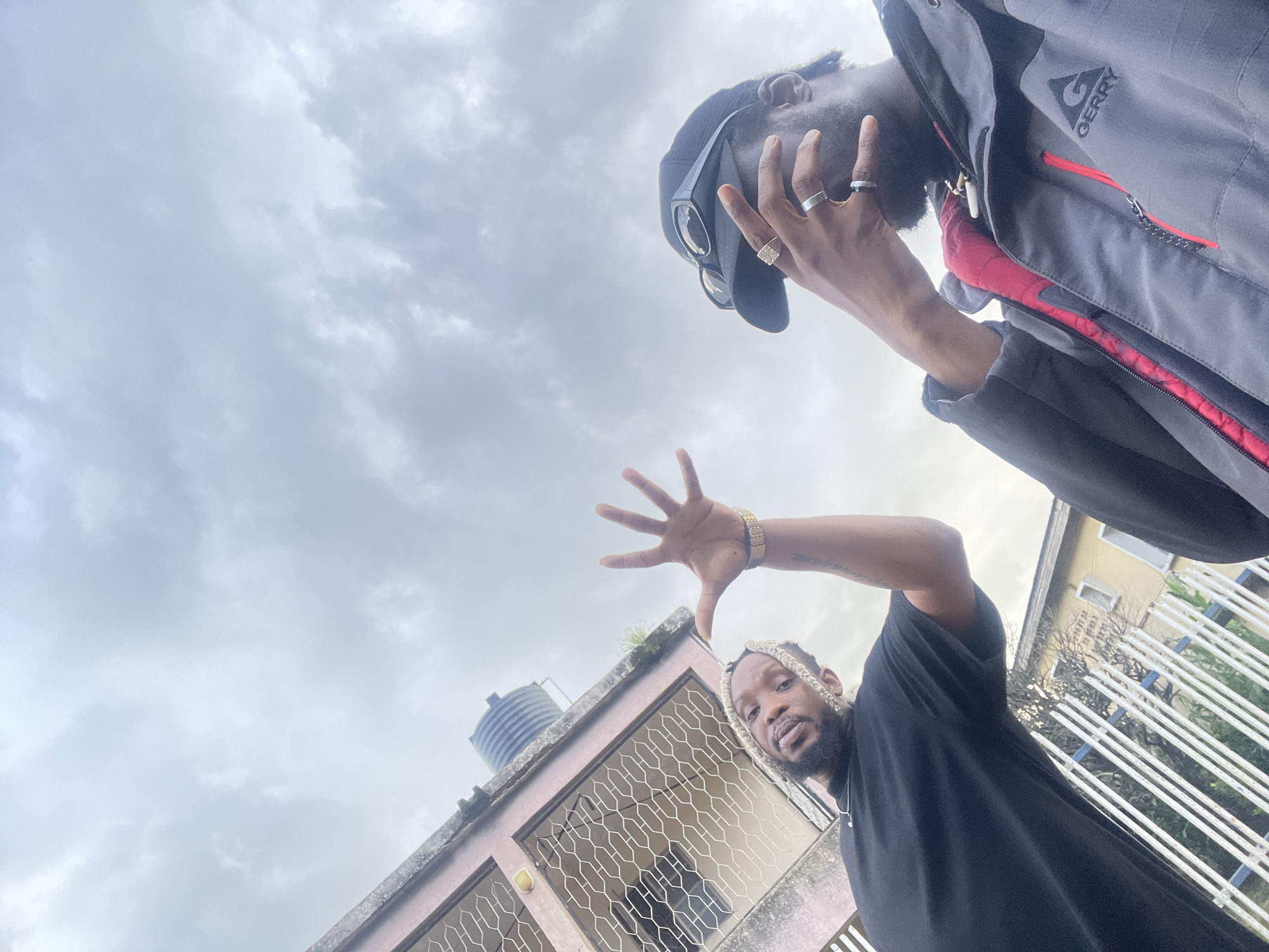 Two men are captured in a low-angle selfie taken outdoors in Jos, Nigeria. The sky above is overcast with textured clouds. On the left, a man with blonde braided hair wears a black t-shirt and a gold watch. He extends his left hand, palm facing the camera, with fingers spread. To his right, a man in a black baseball cap, sunglasses, and a grey and red jacket covers his face with his right hand, revealing multiple rings on his fingers. Behind the men, a pink building with barred windows is visible, topped by a water tank. A white metal fence runs across the foreground, partially obscuring a yellow building in the background.