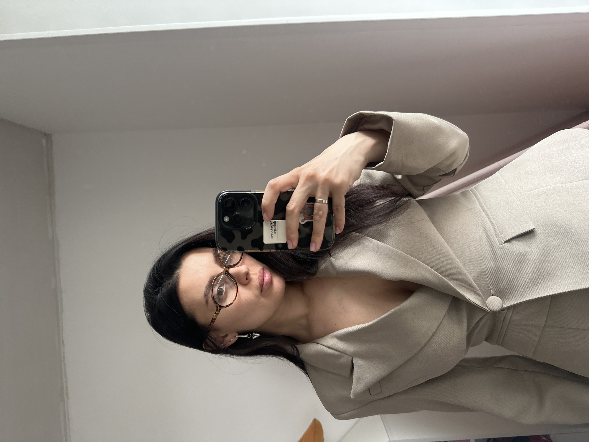 A woman stands indoors, holding a cell phone in her right hand to take a selfie. She is wearing glasses with tortoiseshell frames and an outfit that appears to be a beige blazer dress with a single button closure. The location is likely indoors, based on the presence of interior walls and what appears to be a mirror in the background. There is no clear indication in the image that allows to confirm it has been taken in the city of Chaikovsky, Russia.