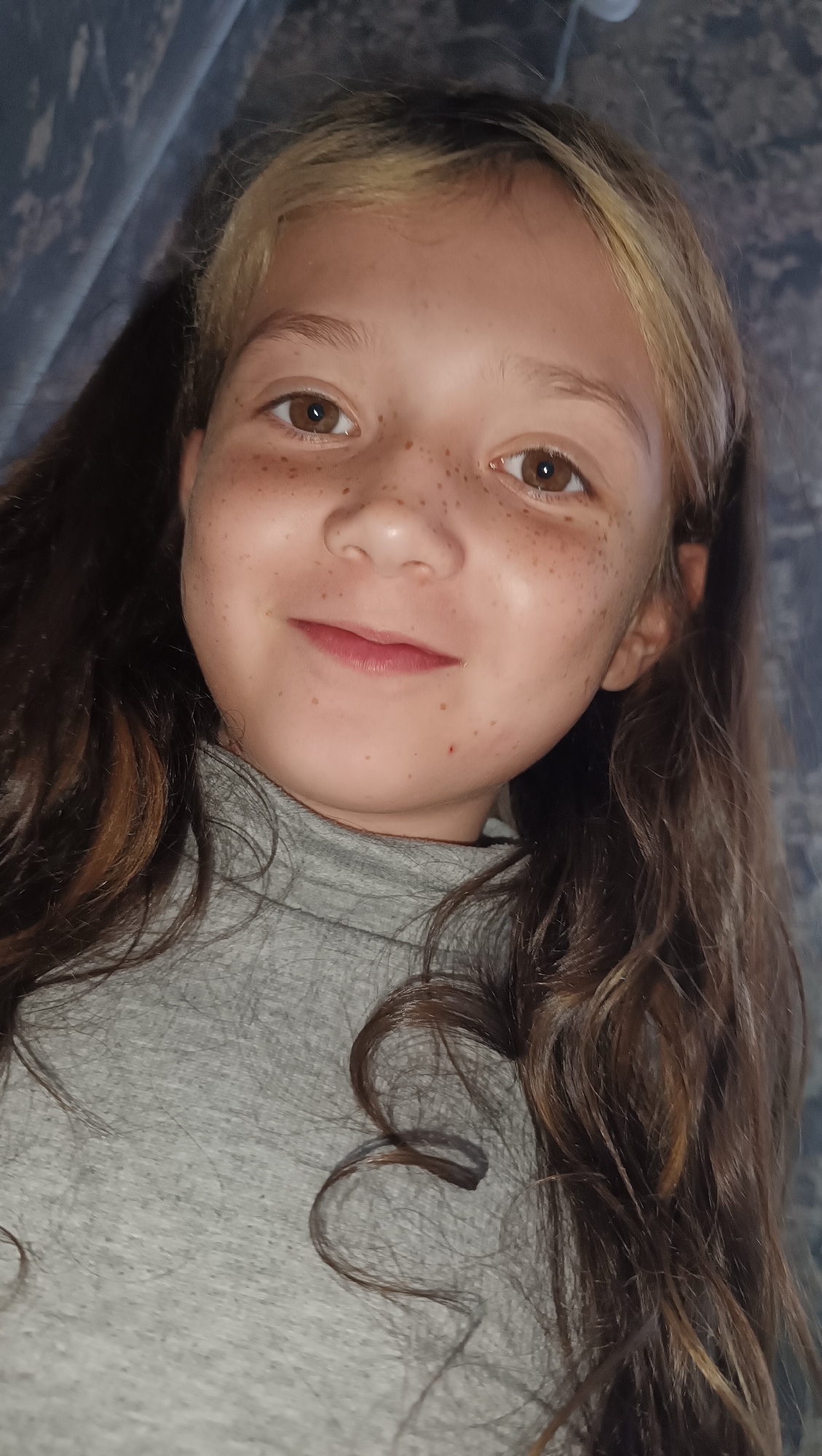 The image is a close-up, vertical, selfie-style photograph of a young girl with light brown hair and brown eyes. She has a light dusting of freckles across her nose and cheeks. Her hair is long and wavy, with some lighter streaks. She is wearing a gray, high-necked shirt. The background is dark and appears to be a draped fabric or curtain. The lighting suggests an indoor setting, possibly in the evening or with artificial light, and does not provide cues for the weather. The girl's expression is a gentle smile. There is no visible text in the image. The location context provided is Karsha, Russia.