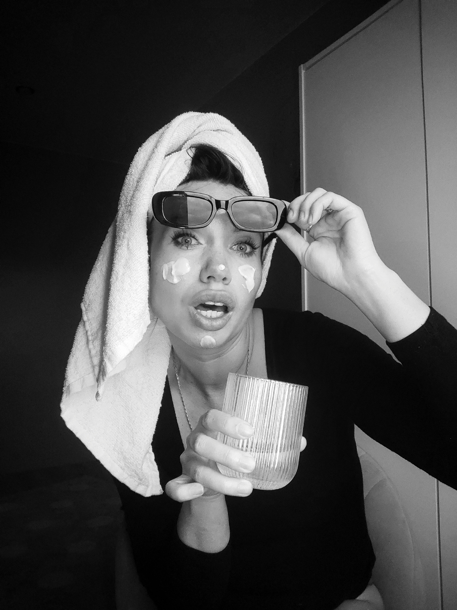 A black and white medium shot captures a woman indoors, possibly in Votkinsk, Russia, engaged in a skincare routine. She has a white towel wrapped around her head, suggesting she has recently washed her hair or face. Her face is dotted with several spots of what appears to be a face cream or mask. She is wearing black, rectangular sunglasses pushed up on her forehead, and her right hand is holding the sunglasses, slightly adjusting them or pulling them further up. Her left hand holds a clear, ridged glass, possibly containing water, which she is bringing towards her body.
The woman's expression is one of surprise or shock, with her mouth agape and her eyes wide, looking directly at the viewer. Her eyebrows are slightly raised. The background is dark and plain on the left, while the right side reveals part of a light-colored wall or door, indicating an indoor setting. There are no specific cues for the time of day or weather. No visible text is present in the image. The overall impression is candid and somewhat humorous, capturing a moment of unexpected interruption during a personal grooming routine.