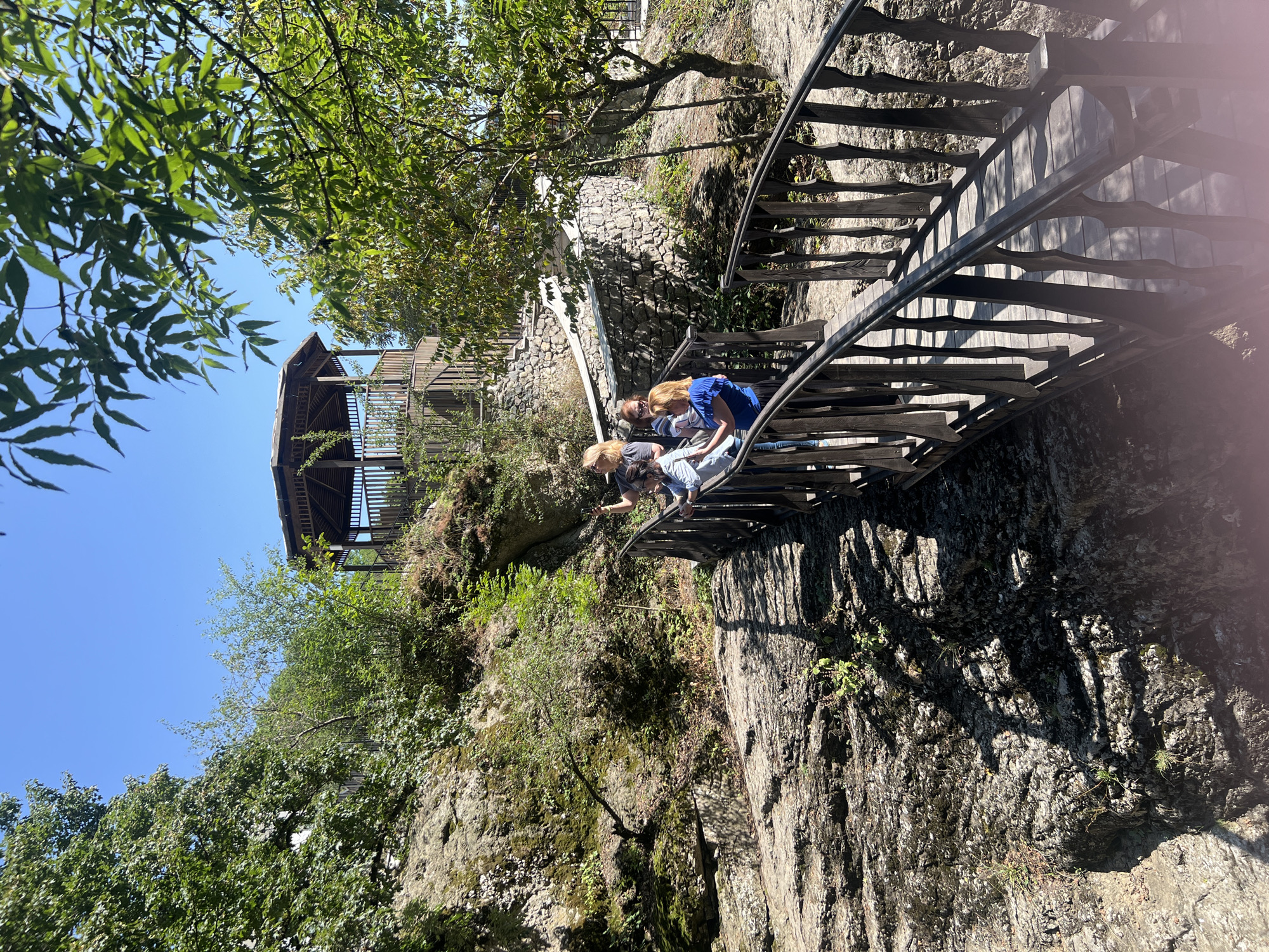 A group of four people, three women and one child, are walking across a wooden bridge in a rocky, forested area. The bridge has an ornate railing. In the background, a wooden gazebo with a sloped roof sits on a hill. The sky is clear and blue. This scene is located in the vicinity of the city of Sarısa, Azerbaijan.