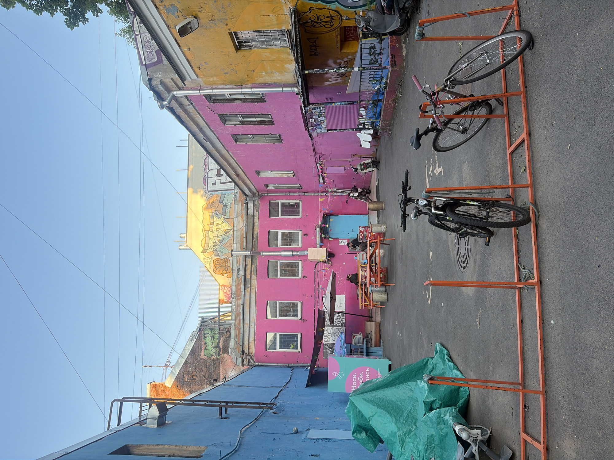 This is a medium shot of an outdoor courtyard area in Saint Petersburg, Russia. The courtyard is enclosed by brightly colored buildings; the left building is painted bright blue, the central building is painted hot pink, and the right building is painted bright yellow. The buildings have multiple windows and are adorned with graffiti. In the foreground, there is an orange metal bicycle rack holding two bicycles. A green tarp covers an object on the left side of the courtyard. In the middle ground, there are picnic tables with people seated at them. Several motorcycles and scooters are parked throughout the courtyard. Overhead, power lines crisscross the clear blue sky. The overall impression is of an urban, artistic, and somewhat weathered space.