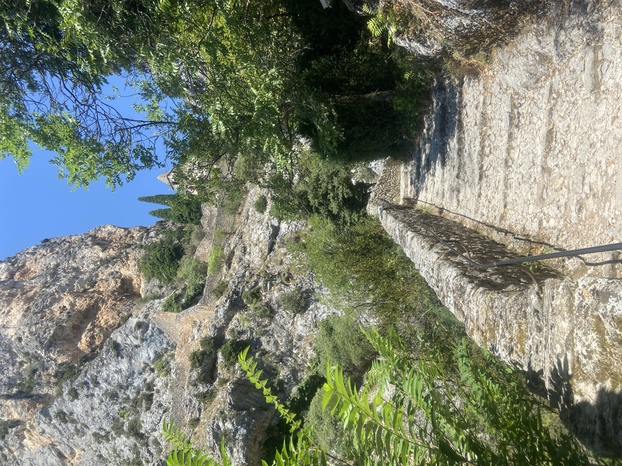 A stone staircase winds its way up a rocky mountainside, flanked by a low stone wall on the left and dense foliage on the right. The staircase is made of uneven, weathered stones, suggesting an ancient path. Above and to the right of the staircase, a stone building with a steeple is visible among the trees. The mountainside is steep and rugged, with visible rock formations and patches of greenery. The sky is a clear blue, indicating a sunny day. The overall scene depicts a natural, historical, and potentially religious site.