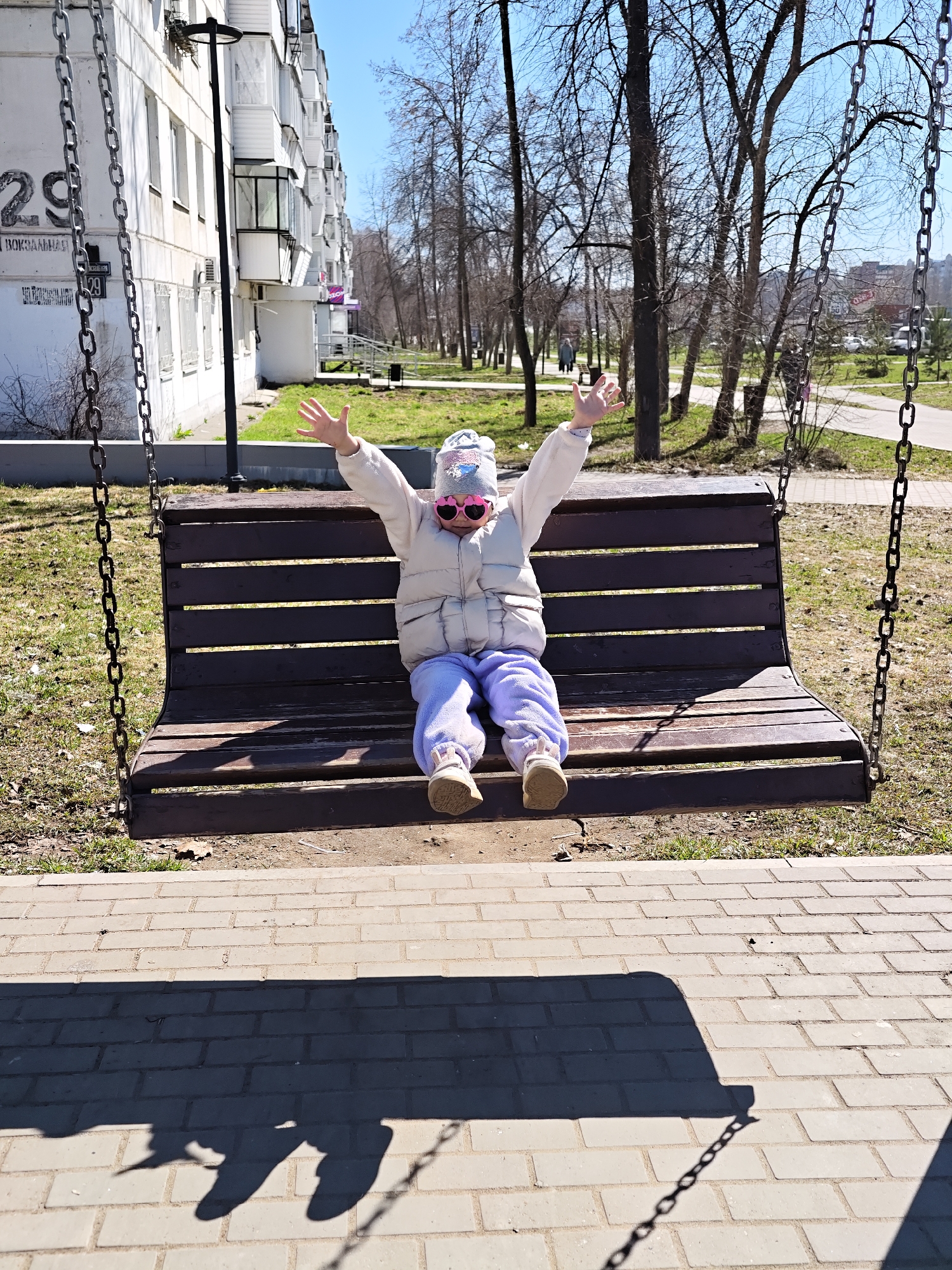 A young child wearing a white puffy vest, lavender sweatpants, and heart-shaped pink sunglasses sits on a wooden swing in a city park in Chaikovsky, Russia. The child has their arms raised in apparent enjoyment. The swing is suspended by metal chains. Behind the swing is a grassy area with trees and a pedestrian walkway. In the background, there is an apartment building with a sign that reads "29 Вокзальная". The ground in front of the swing is paved with grey bricks, on which the shadow of the swing is cast. The day appears to be sunny.