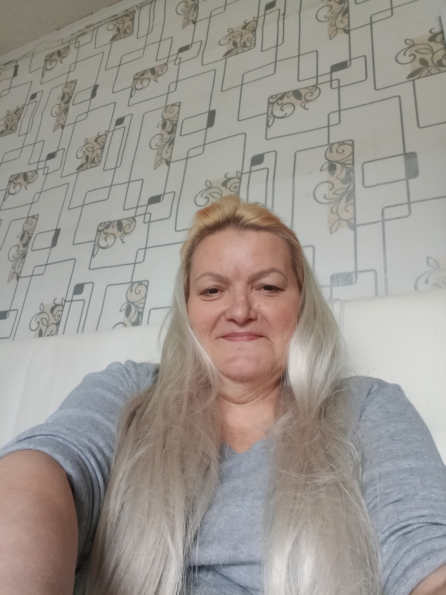 A woman with blond hair pulled into a bun and long strands falling around her face is sitting on a white surface, likely a bed or couch. She is wearing a gray, long-sleeved shirt. The background is a wall with a patterned wallpaper, featuring geometric shapes and floral designs. The image was taken indoors. There is no information suggesting this image was taken in the city of Красный Маяк, Russia.