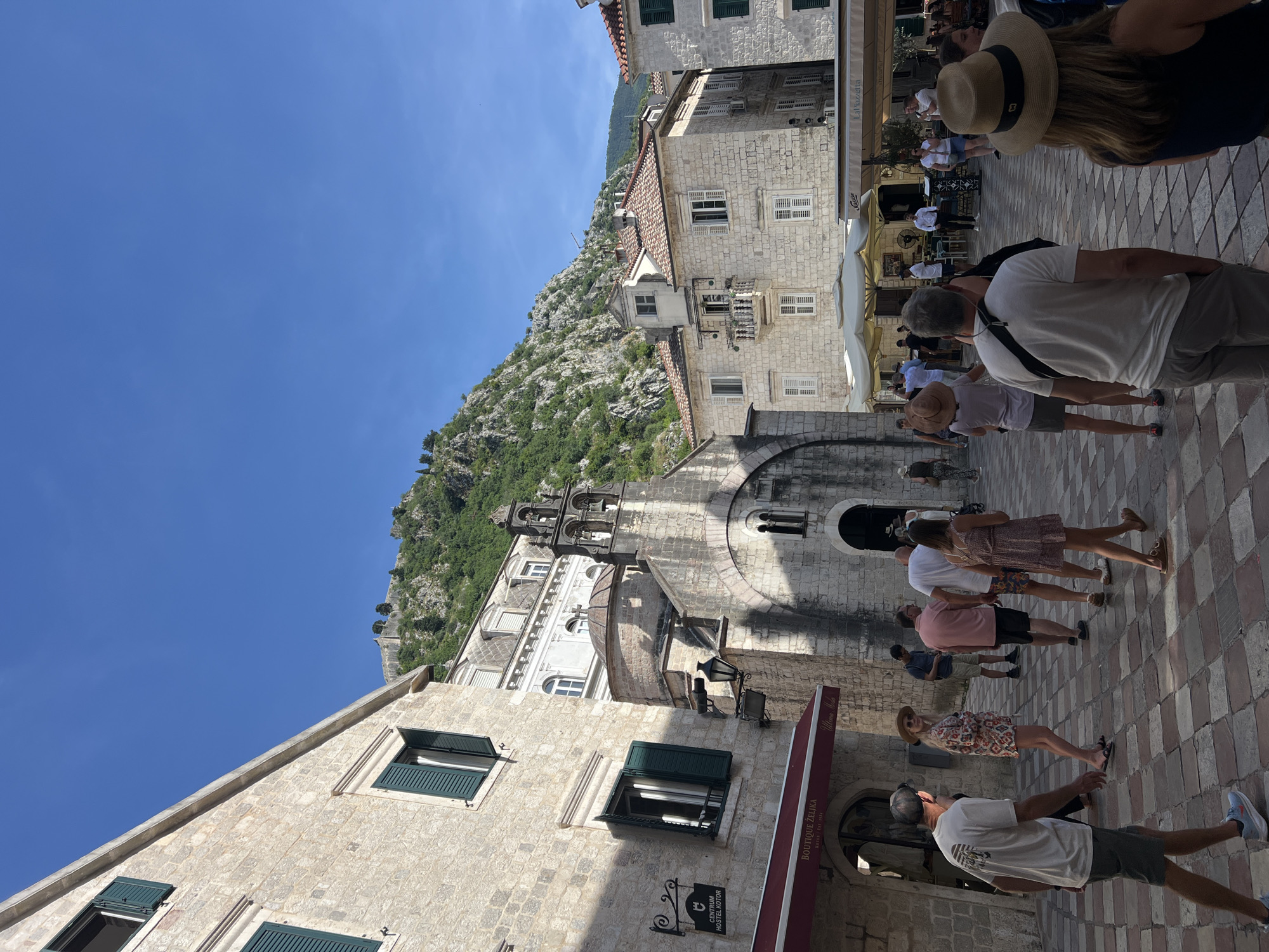 This detailed image depicts a bustling stone-paved square within the Old Town of Kotor, Montenegro, identifiable by the "CENTRUM HOSTEL KOTOR" sign and the distinct mountainous backdrop featuring the St. John's Fortress walls.
In the foreground, numerous individuals, approximately twenty, populate the square, walking or standing. They are casually dressed in summer attire, including shorts, t-shirts, dresses, and hats. The paving consists of light and dark checkered stone tiles.
To the left, a multi-story stone building features several windows with open green shutters and a red awning over "Boutique ŽELJKA." Below one window, a black wrought-iron sign reads "CENTRUM HOSTEL KOTOR." Further back, a smaller stone church with a central dome, a prominent bell tower containing two bells, and an arched entrance is visible. Two ornate black street lamps are affixed to the church's exterior.
Behind the church, a white building with an ornate facade and numerous windows is partially visible. To the right of the church, another stone building with a red tiled roof, featuring a small balcony and multiple windows with white shutters, stands alongside other structures. Several white and beige outdoor umbrellas mark the presence of a café or restaurant, with a sign visible for "La Piazzetta."
The background is dominated by a steep, rugged mountain covered in green foliage and visible rock faces. Near the summit, ancient fortification walls and structures are discernible against a clear, vibrant blue sky. No clouds are present.
