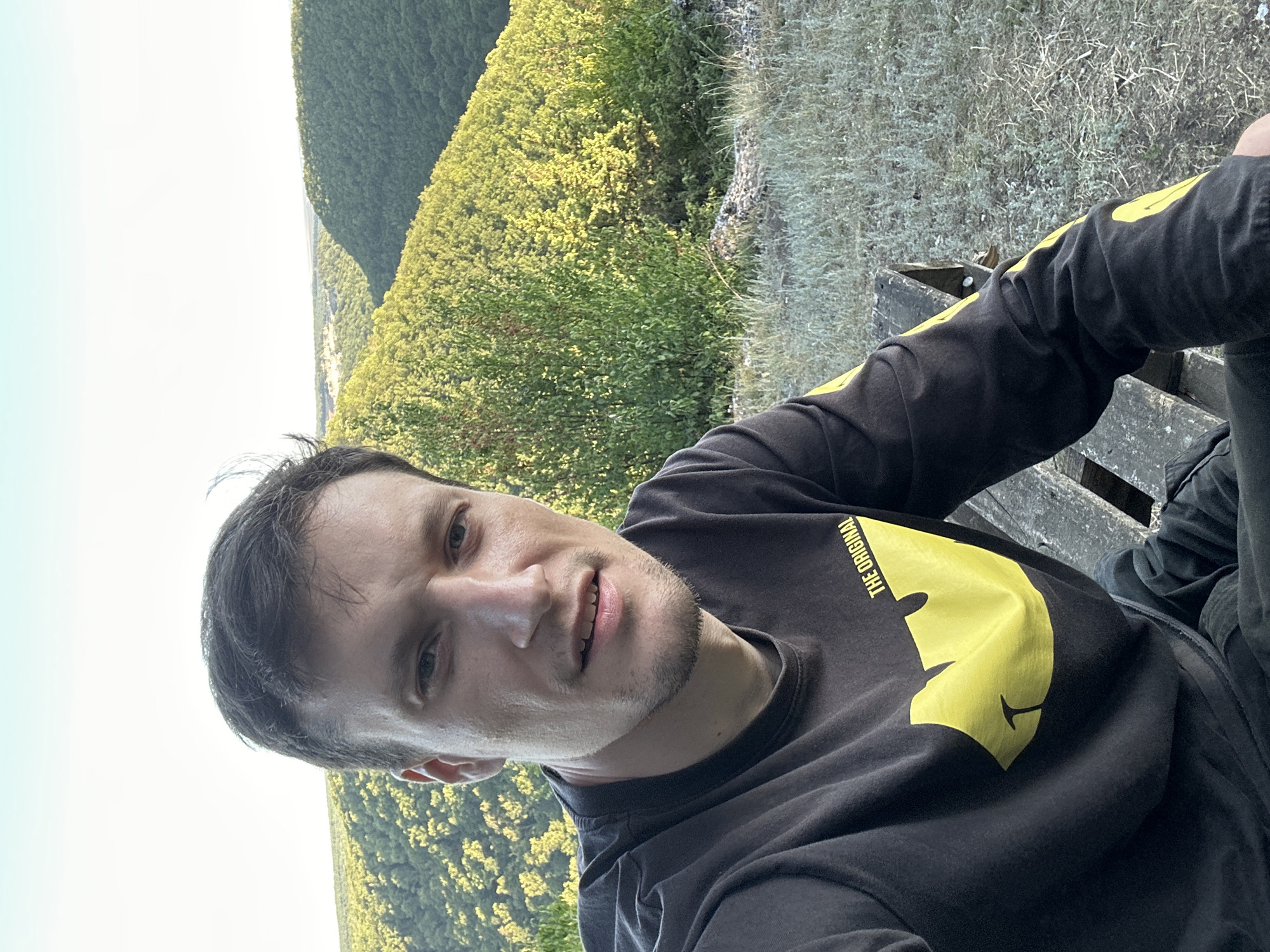 A young adult male with short, dark hair and stubble is captured in a selfie outdoors. He is wearing a black long-sleeved shirt with a yellow Wu-Tang Clan logo and text that reads "THE ORIGINAL" on the left chest. The man is seated on a rustic wooden bench, which is partially visible in the foreground. He is looking directly at the camera with a neutral expression.
The background reveals a picturesque natural landscape characterized by lush green, tree-covered hills and valleys, suggesting a rural or park-like setting. The sky above is a clear, pale blue, indicating daytime. The overall impression is one of a casual outdoor moment, possibly during a hike or a visit to a scenic overlook. The location is identified as Pryvorottia Druhe, Ukraine, a village known for its natural beauty and rural environment. The time of day appears to be late afternoon or early evening, given the soft lighting that casts subtle shadows.