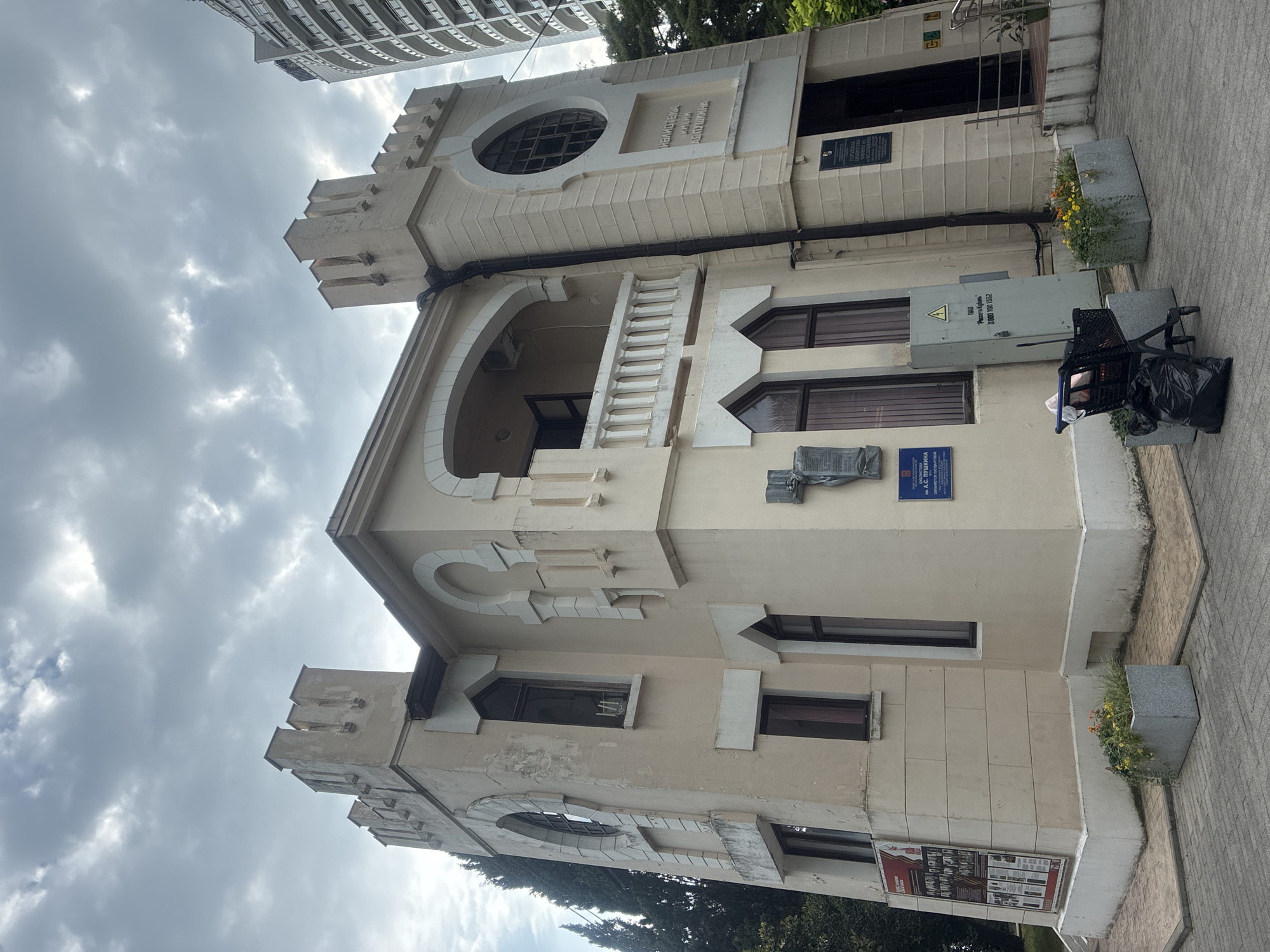 A two-story, cream-colored building with turret-like structures on its corners stands prominently under a cloudy sky in Sochi, Russia. The building's facade features arched windows and decorative elements. To the right of the main building, a portion of a modern, multi-story residential building is visible. In front of the cream-colored building, there is a paved walkway with two planters containing flowers and greenery. A shopping cart with a black trash bag is positioned near the planters. Various signs are affixed to the building's exterior, including what appears to be a historical marker and a blue plaque with text. The overall scene suggests an urban public space, possibly a historical site or a cultural institution.