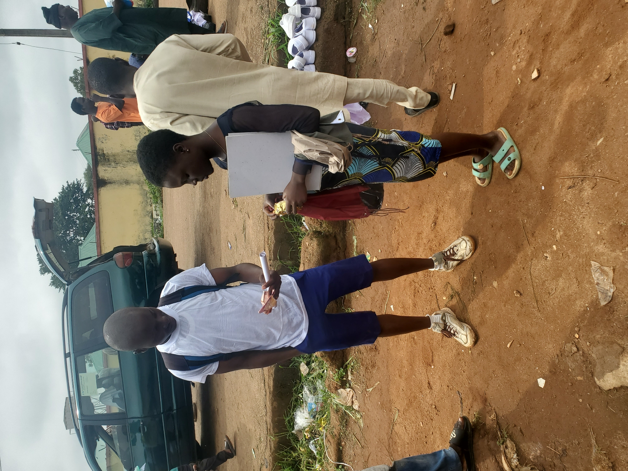 In Bahit, Nigeria, a young Black male wearing a white t-shirt, blue shorts, and white sneakers stands with his hands in his pockets, looking down at some banknotes he is holding. Beside him, a young Black female, dressed in a dark top with sheer sleeves and a patterned skirt, holds a white folder and a red bag. In the background, a green van with its rear hatch open is parked. Several other people are visible further back, some wearing traditional Nigerian attire. The ground is dirt, with scattered debris including plastic bags and wrappers. A wall and some trees are visible in the distance under an overcast sky. A pile of white shoes is arranged on the ground to the right of the main subjects.