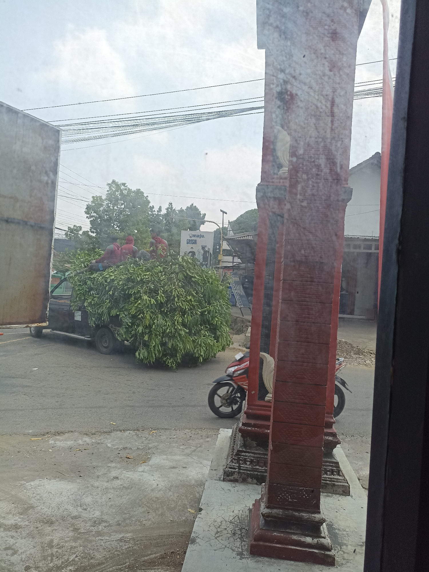 In the city of Pulisen, Indonesia, a dark-colored pickup truck is heavily laden with a large quantity of green foliage, obscuring most of its body. Three individuals, wearing red shirts, are positioned on top of the foliage, appearing to be securing or arranging it. The truck is on a paved road, which is somewhat worn and shows signs of patching. In the background, there are trees, buildings, power lines, and a billboard with text that includes "MAJU. GROSIR". To the right foreground, a red motorcycle is parked, partially visible behind a red, textured pillar. The overall environment suggests an outdoor, possibly urban or semi-urban, setting with overcast skies.