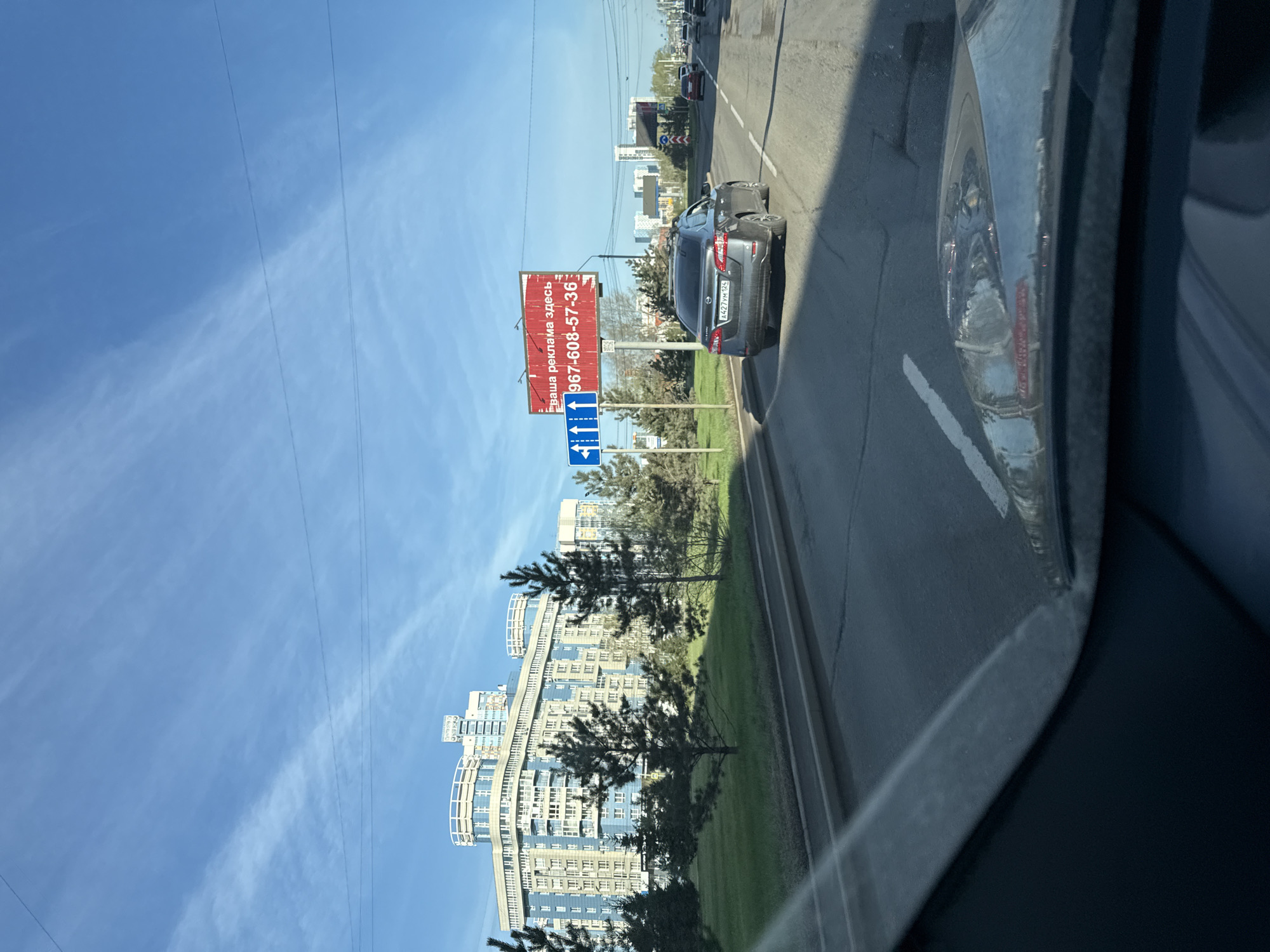 The image shows a view from inside a vehicle traveling on a road in Krasnoyarsk, Russia. A gray Nissan Sentra is driving in the adjacent lane. A red and white advertising billboard displaying the Cyrillic text "Ваша реклама здесь +967-608-57-36" is visible. Further ahead, there are other vehicles on the road and buildings in the distance. A tall, modern residential building with a curved facade is also visible. The sky is blue with thin clouds.
