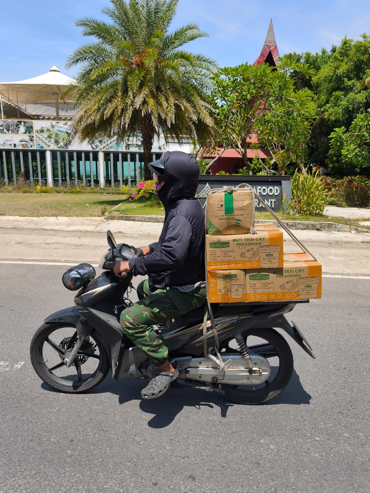A man wearing a black hoodie and camouflage pants rides a black Honda motorcycle on a paved road. He is wearing a black cap and sunglasses, and the hoodie is pulled up to cover his face and neck. The motorcycle is carrying several boxes stacked on the rear rack, secured with rope. The boxes appear to contain food products, with labels indicating "GOLDEN FARM PRESERVES." In the background, there is a palm tree, a building with a white canopy, and a restaurant sign. The sky is blue and clear, suggesting a sunny day. The location is identified as Bac Nha Trang Ward, Vietnam.