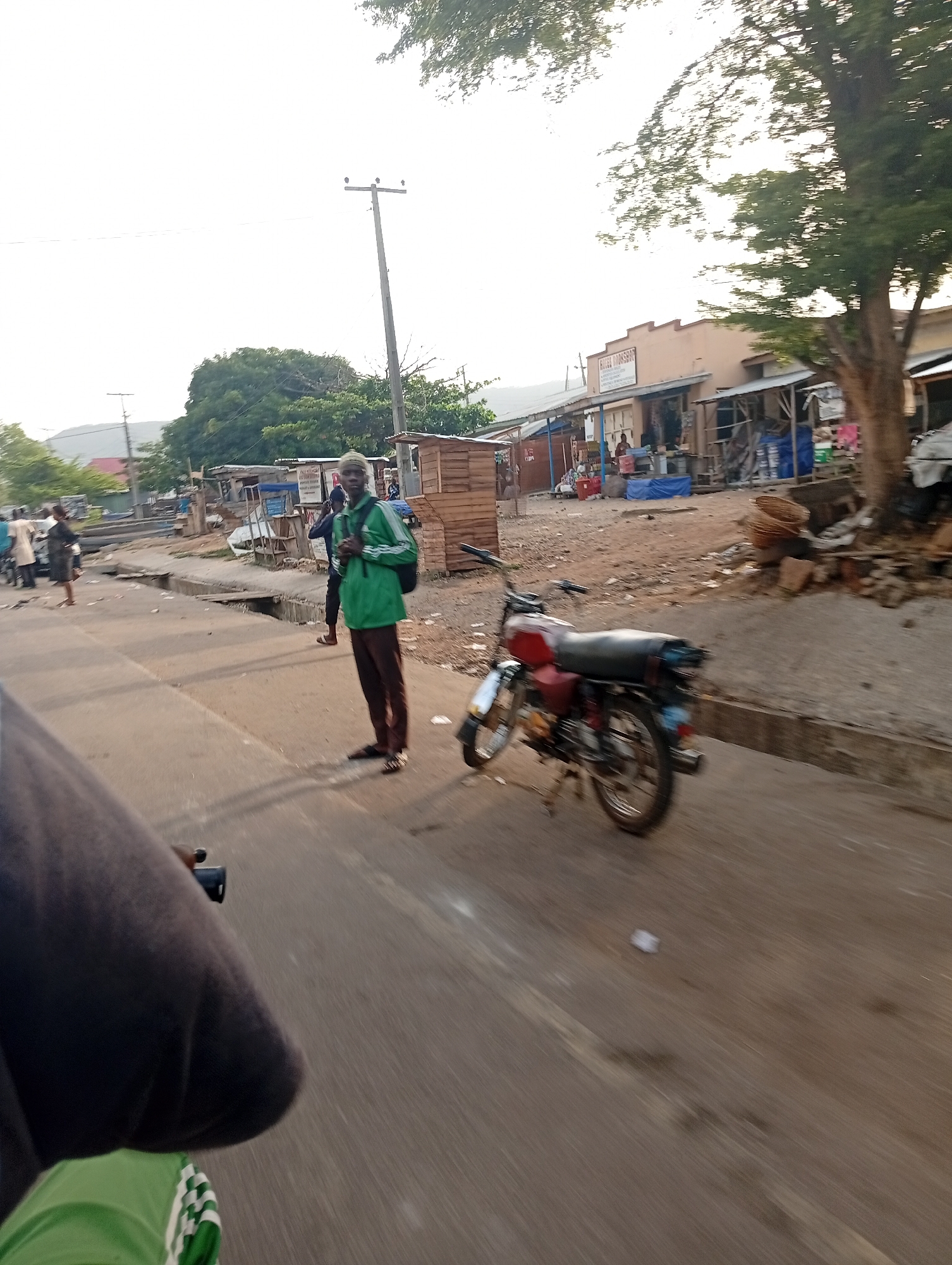 A street scene in Adankolo, Nigeria. A man wearing a green jacket and brown pants stands on a concrete sidewalk, near a motorcycle parked on the edge of the road. In the background, there are several small shops and people standing around. Power lines are visible overhead, and there are trees on both sides of the road. The road is paved and appears to be in a residential area.