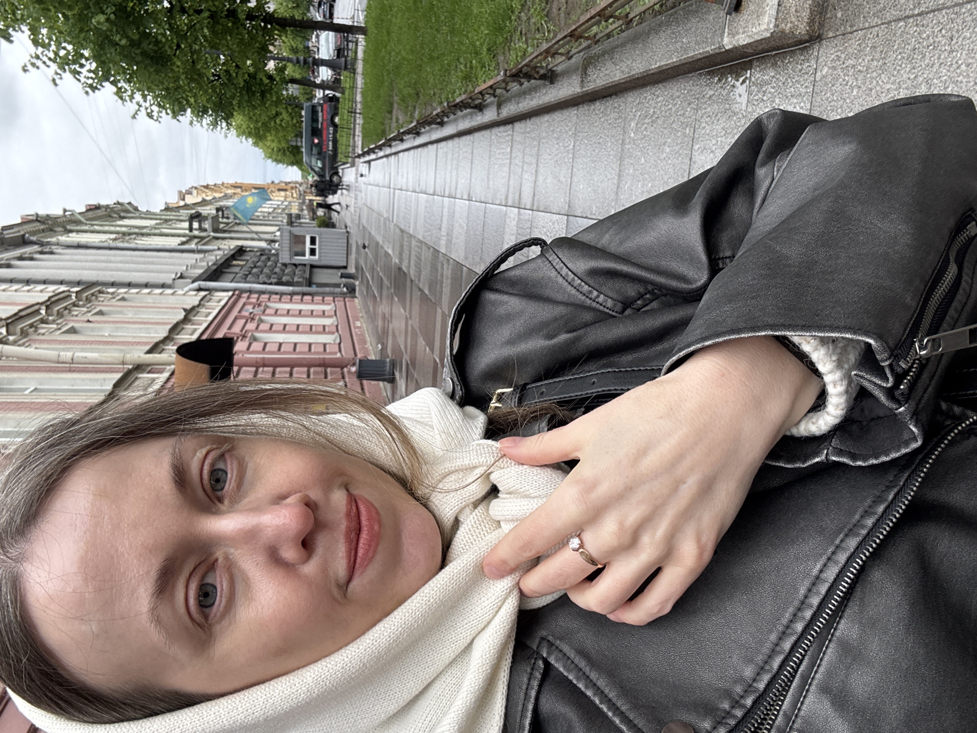 A selfie shows a woman on a street in Saint Petersburg, Russia. She has light skin, grey hair, and blue eyes. She wears a black leather jacket over a white knit scarf. On her ring finger, there is a ring with a clear stone.
Behind her, a wet, grey sidewalk extends along a street lined with old buildings. Some are pink and white, others white or grey. Trees line the street and several vehicles are parked along the curb. A flag hangs from one of the buildings. A guardhouse with a person visible in it is also seen.