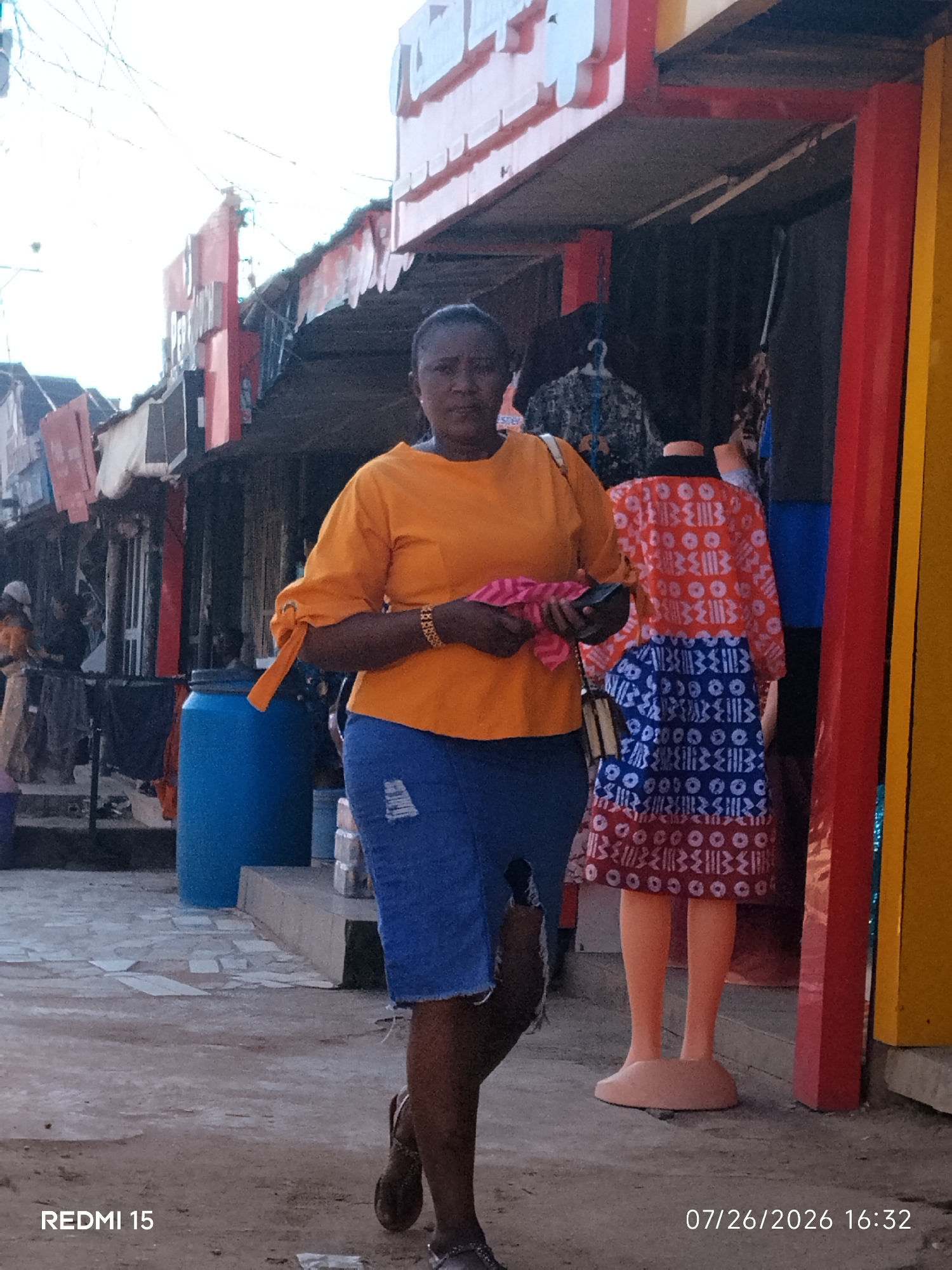 In Umuode, Nigeria, a woman walks on a paved area in front of shops. She is wearing an orange short-sleeved shirt with ruffled details on the sleeves and blue denim shorts. In her left hand, she holds a pink folded item and a dark clutch purse. She is also wearing a gold bracelet on her left wrist and a necklace. A mannequin wearing a vibrant orange and blue patterned dress stands to her right. The storefronts display various items, and a large blue barrel is visible to the left of the woman. The image is dated 07/26/2026 at 16:32.
