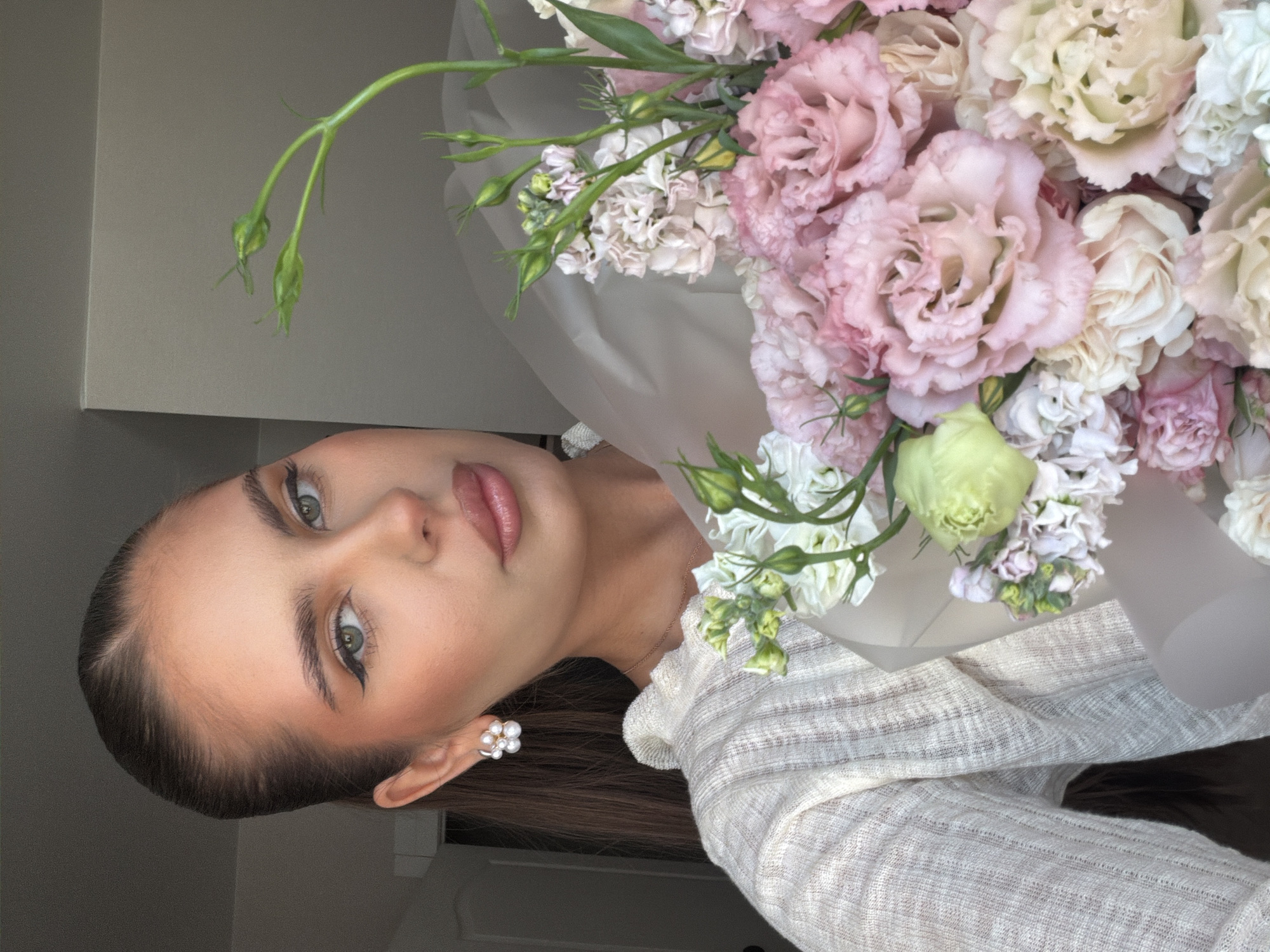 A woman poses with a large bouquet of pink and white flowers, possibly lisianthus. She is wearing a light-colored, textured top and pearl earrings. Her hair is dark and pulled back. She has light skin, green eyes, and is wearing eyeliner. The background is a plain, light-colored wall. There is no indication of this photo being taken in Krasnoyarsk, Russia.