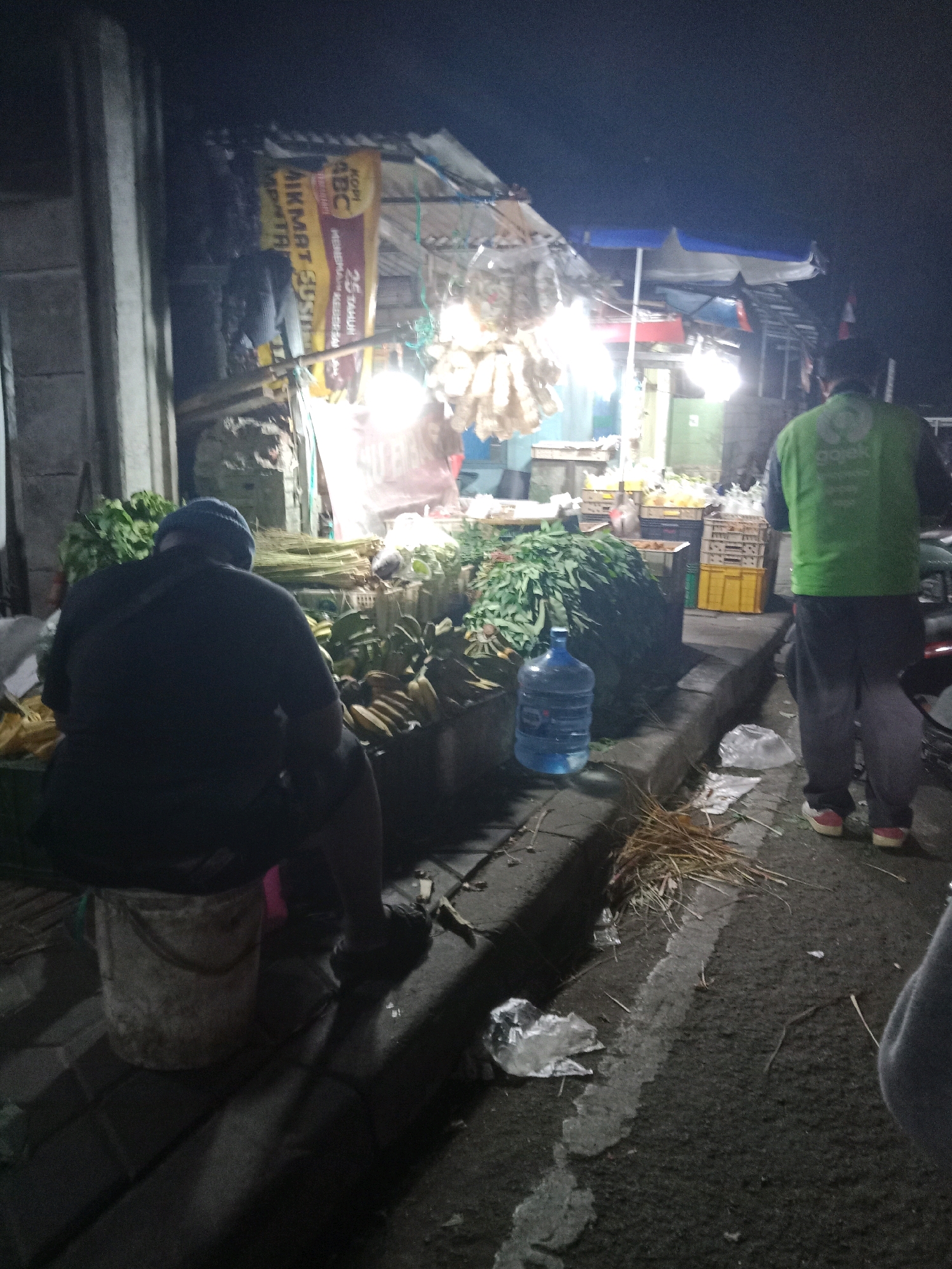 A low-light, slightly blurry nighttime street scene in South Tangerang, Indonesia, depicts a market stall. In the foreground, a person wearing a dark jacket and a blue beanie sits on a white bucket, their back to the camera. To their right, a large blue water bottle stands near a pile of produce. Further back, another person, wearing a bright green vest with a logo that appears to be "Gojek," stands facing away. The stall itself is laden with various fresh produce, including what appears to be bananas and leafy green vegetables, some arranged in crates. Overhead, lights illuminate the scene, casting shadows and highlighting the market atmosphere. A yellow banner with Indonesian text, including "ABC" and "AIKMAT SUSI," is visible in the background. The ground is paved, and debris, including straw and plastic, is scattered on the asphalt. The overall impression is that of a bustling, albeit dimly lit, nighttime market.