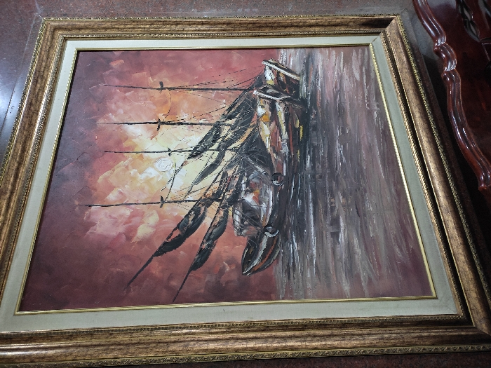 The image displays a framed oil painting, likely situated indoors against a dark, possibly reddish-brown wall. A dark, polished wooden object, possibly furniture, is partially visible in the bottom right foreground.
The painting itself, rendered in an impasto style, depicts a maritime scene. Three traditional sailing or fishing boats with tall, dark masts and partially furled, dark sails are positioned on a body of water. The boats are dark-toned, predominantly brown and black, with some lighter accents on their hulls. The water, rendered with visible brushstrokes, appears choppy and is depicted in dark grey and brown hues. The sky is a dramatic expanse of thick, textured brushstrokes in shades of deep red, orange, and yellow, suggestive of a sunrise or sunset. A faint, circular light source, resembling the sun or moon, is visible in the upper-center portion of the sky.
The painting is enclosed within an ornate, multi-layered frame. The outermost frame is wide, gilded, and features a textured or carved pattern. Inside this, a narrower, light beige or off-white mat separates the gilded frame from a thin, plain gold inner lip that directly borders the artwork. No people, specific actions, or interactions are discernible in the scene. The precise location within Pacet, Indonesia, cannot be determined from the visual information provided in the image.