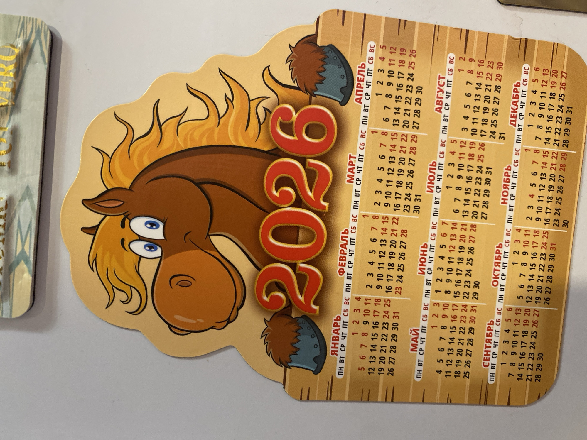 This image depicts a wall calendar for the year 2026. The calendar features a cartoon illustration of a horse's head and mane at the top, with the numbers "2026" prominently displayed below the horse. The calendar is divided into months, with each month's name written in Russian. The days of the week are also abbreviated in Russian. The calendar is attached to a plain, light-colored wall. There is no discernible information in the image that would suggest its location in Novaya Usman', Russia, or any other specific city.