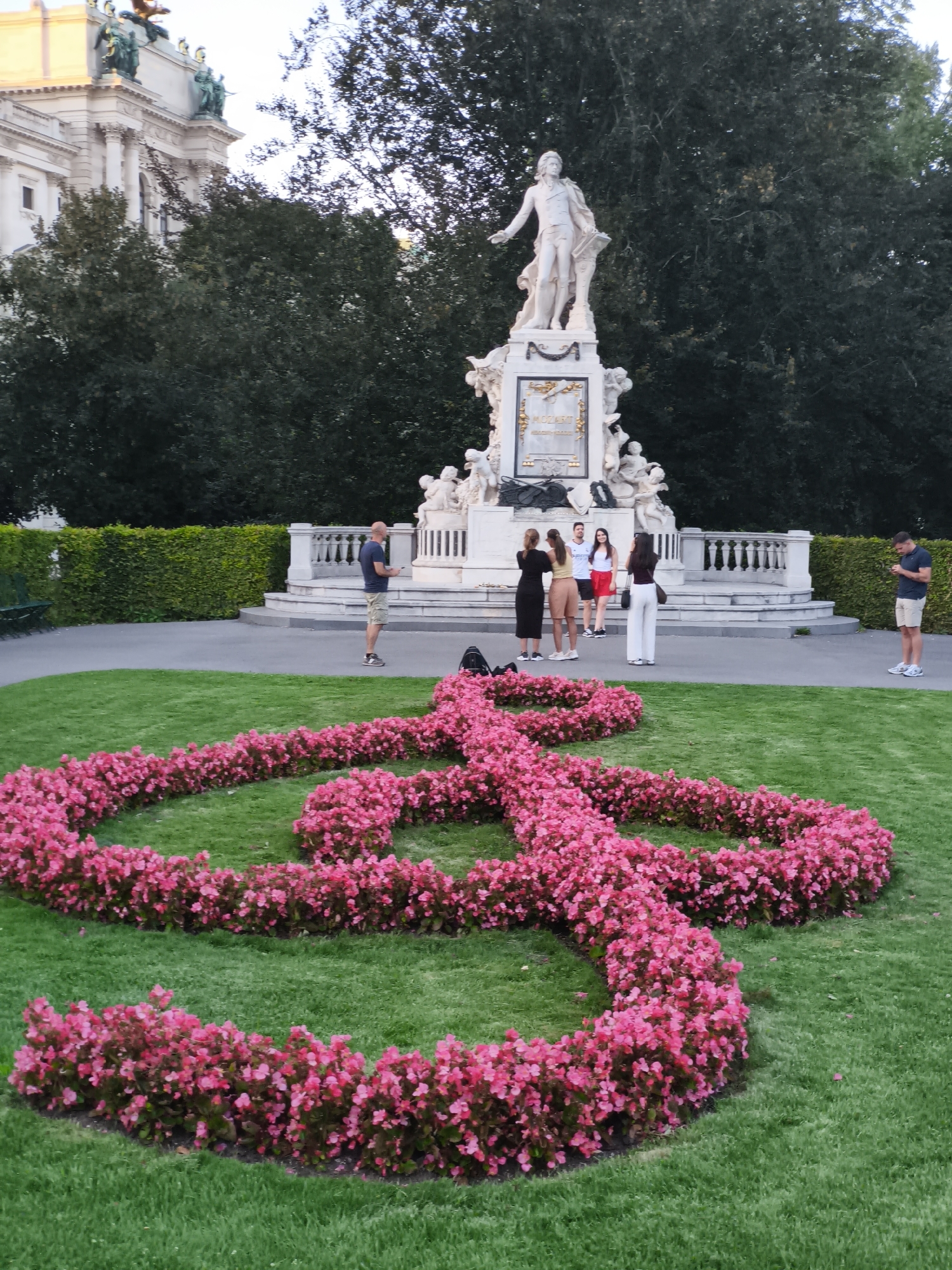 The image depicts the Mozart Monument in Burggarten, Vienna, Austria. The monument is a grand marble structure featuring a statue of Wolfgang Amadeus Mozart and surrounding allegorical figures. In the foreground, a large, heart-shaped arrangement of bright pink flowers is laid out on a meticulously maintained green lawn. Several people are visible near the monument. A man in a blue shirt and shorts stands to the left, looking at his phone. A group of four individuals, two men and two women, are gathered near the base of the monument. Another man in a blue shirt and shorts is on the right side of the frame, also looking at his phone. The background shows lush green trees and a portion of a grand building with classical architectural elements.