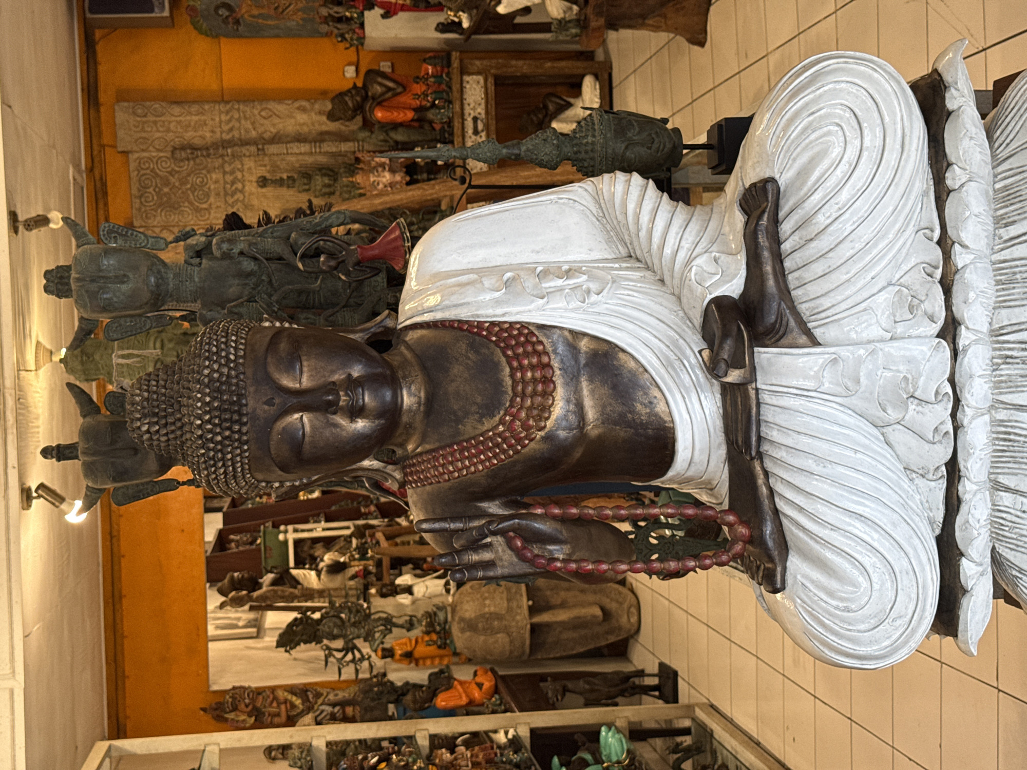 The image prominently features a large, ornate statue of Buddha, presented in a meditative posture, seated cross-legged with hands in the Dhyana Mudra. The Buddha's skin is a dark, metallic bronze color, contrasting sharply with its flowing, textured white robes, which are intricately draped. A strand of reddish-brown mala beads is draped around its neck and held in its clasped hands. The Buddha's face emanates a serene expression, with downcast eyes, and its head is adorned with tightly coiled hair, forming the Ushnisha.
The statue is situated indoors, within what appears to be an artisan shop, gallery, or market stall, fitting the context of Ubud District, Indonesia. The background is densely packed with a variety of other statues and carvings, predominantly in dark, earthy tones of bronze, wood, and stone. Visible among these are numerous smaller Buddha figures, stylized deities, and large carved wooden panels featuring elaborate traditional patterns. Some background figures display hints of orange and red accents. The overall impression is one of a rich collection of spiritual and artistic artifacts.
The scene is illuminated by artificial lighting, including a visible spotlight, highlighting the textures of the displayed items. The floor has light-colored tiles, and reflections suggest the presence of glass display cases or polished surfaces. There is no visible text in the image.