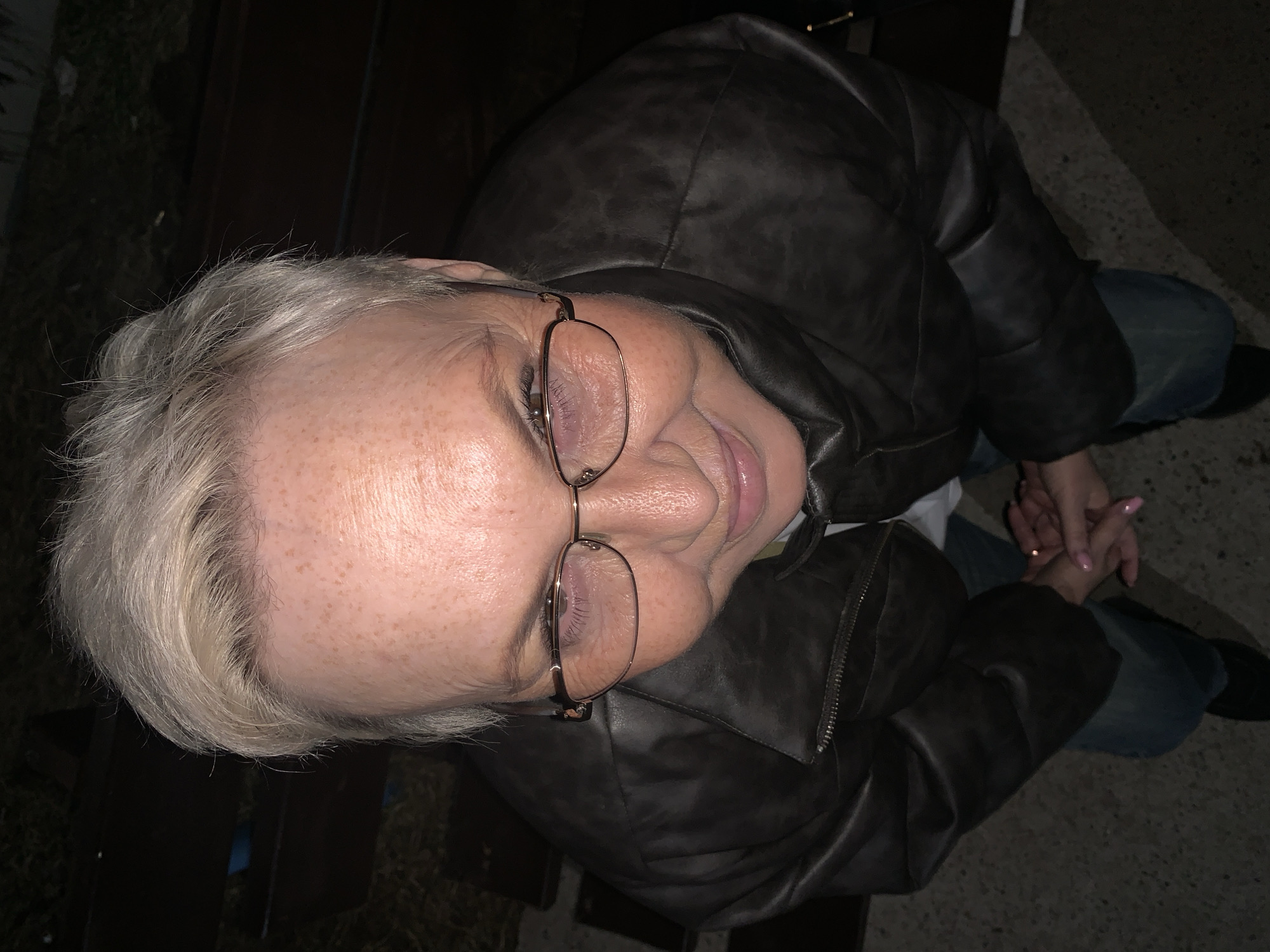 A high-angle view captures an older woman with short, light-grey hair and fair skin speckled with freckles. She wears glasses with thin, gold-toned frames. Her face is tilted upwards. She is smiling slightly. She's wearing a dark brown leather jacket over what appears to be a white shirt. She's also wearing blue jeans and black shoes. Her hands are clasped in her lap, and the fingernails are painted light pink. The person is sitting on a dark wooden bench in what appears to be an outdoor setting. The setting is dark, potentially at night. The specific location in Chaikovsky, Russia, is not discernible from the image.