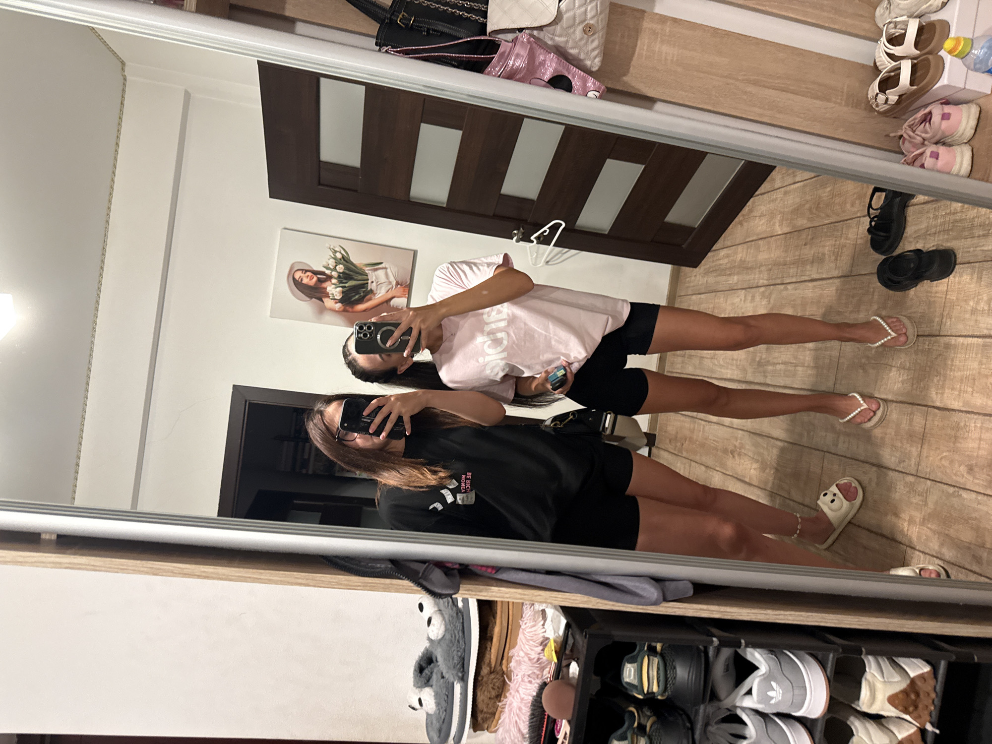 Two young women are taking a mirror selfie in a room that appears to be a closet or dressing area. The woman on the left is wearing a black t-shirt and black shorts, and is holding a phone up to take the picture. She has long brown hair and is wearing glasses. The woman on the right is wearing a pink t-shirt and black shorts, and is also holding a phone. She has dark hair pulled back in a ponytail. They are standing on a wooden floor. To the left of the women is a tall mirror, and to the left of the mirror is a shelf unit filled with shoes. In the reflection of the mirror, a brown wooden door is visible, and on the wall above the door is a framed picture of a woman holding a bouquet of flowers. The lighting in the room is bright, suggesting it is daytime. The overall impression is of a casual, personal space. This scene is likely a private residence in Chișinău, Moldova.