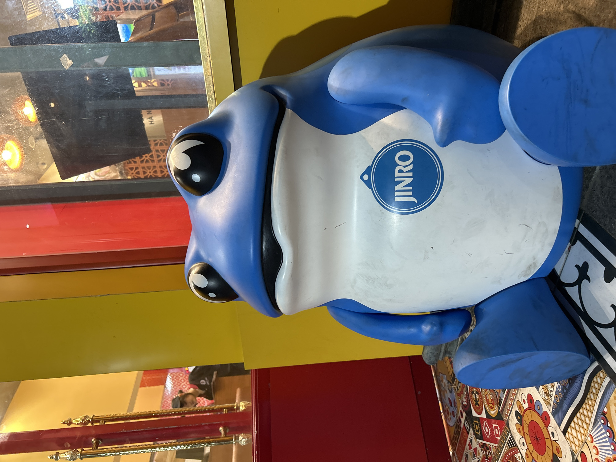 An outdoor scene shows a large, blue cartoon frog statue sitting on the ground. The frog is light blue with white on its belly and has large black eyes with white pupils. A circular blue logo with the word "JINRO" in white letters is visible on the frog's chest. The frog is positioned in front of a building with red and yellow walls and a window. The ground is covered with a patterned tile floor. In the background, through the window, a person wearing a black uniform and a cap is visible. The overall scene appears to be located in a commercial area, possibly a street or entrance to a business.