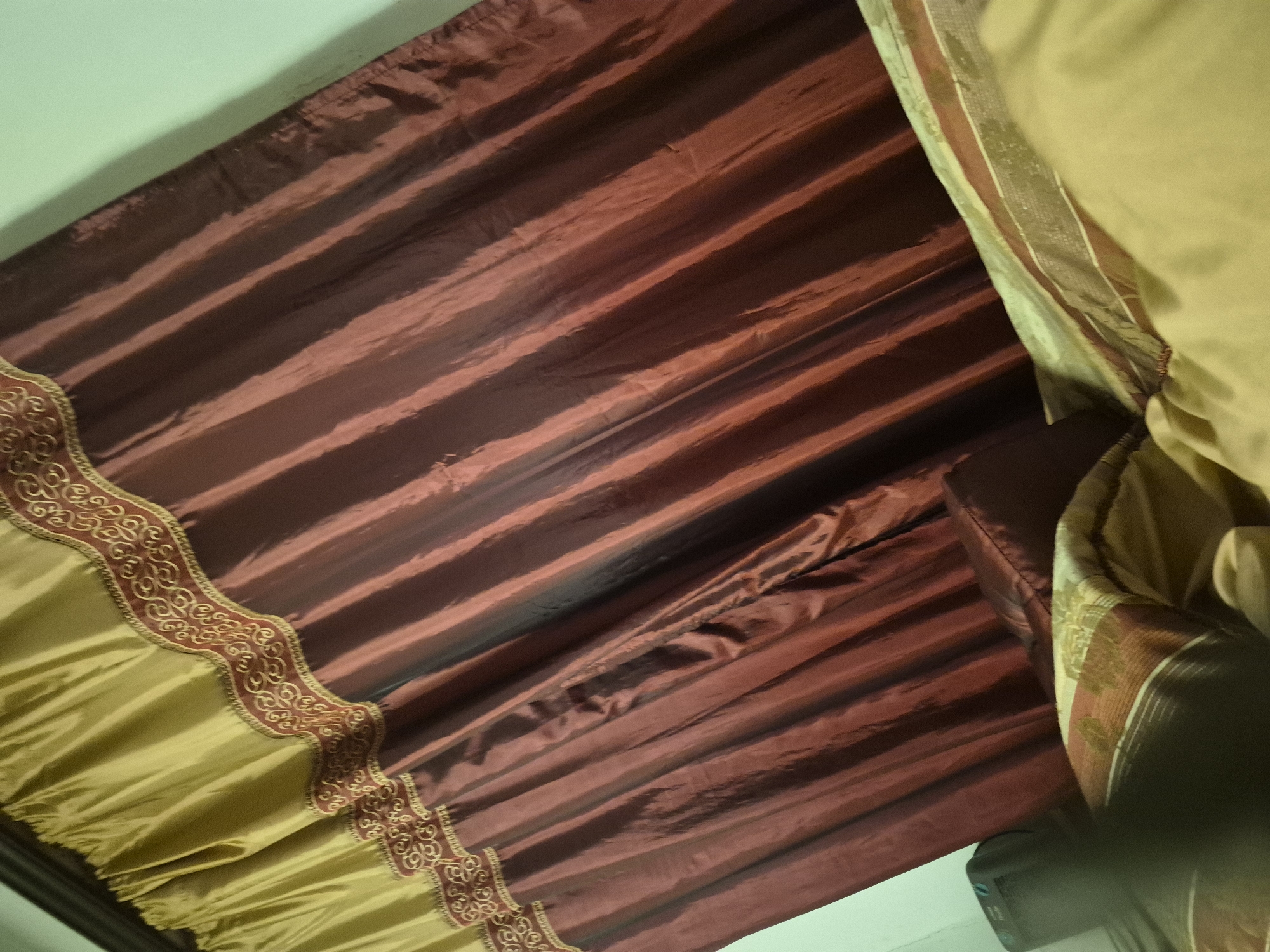 This image depicts an interior scene, likely a room within a residence in Vanadzor, Armenia. The dominant feature is a set of curtains covering a window, captured from a slightly upward and diagonal perspective. The top section of the curtains is fabricated from a gathered, golden-tan material, featuring an ornate embroidered border with swirling gold patterns on a maroon background. Below this border, the main body of the curtains is a solid, deep maroon fabric, exhibiting distinct vertical folds and a subtle sheen. A light mint-green wall is visible to the upper right of the curtains. In the lower foreground, a bed or sofa is partially visible, covered with a golden-yellow sheet and a patterned duvet or blanket featuring maroon, brown, and light-colored stripes and motifs. A maroon pillow is also present on the bed/sofa. A dark, rectangular electronic device is partially visible on the far lower-left side of the frame. No individuals are present in the image.