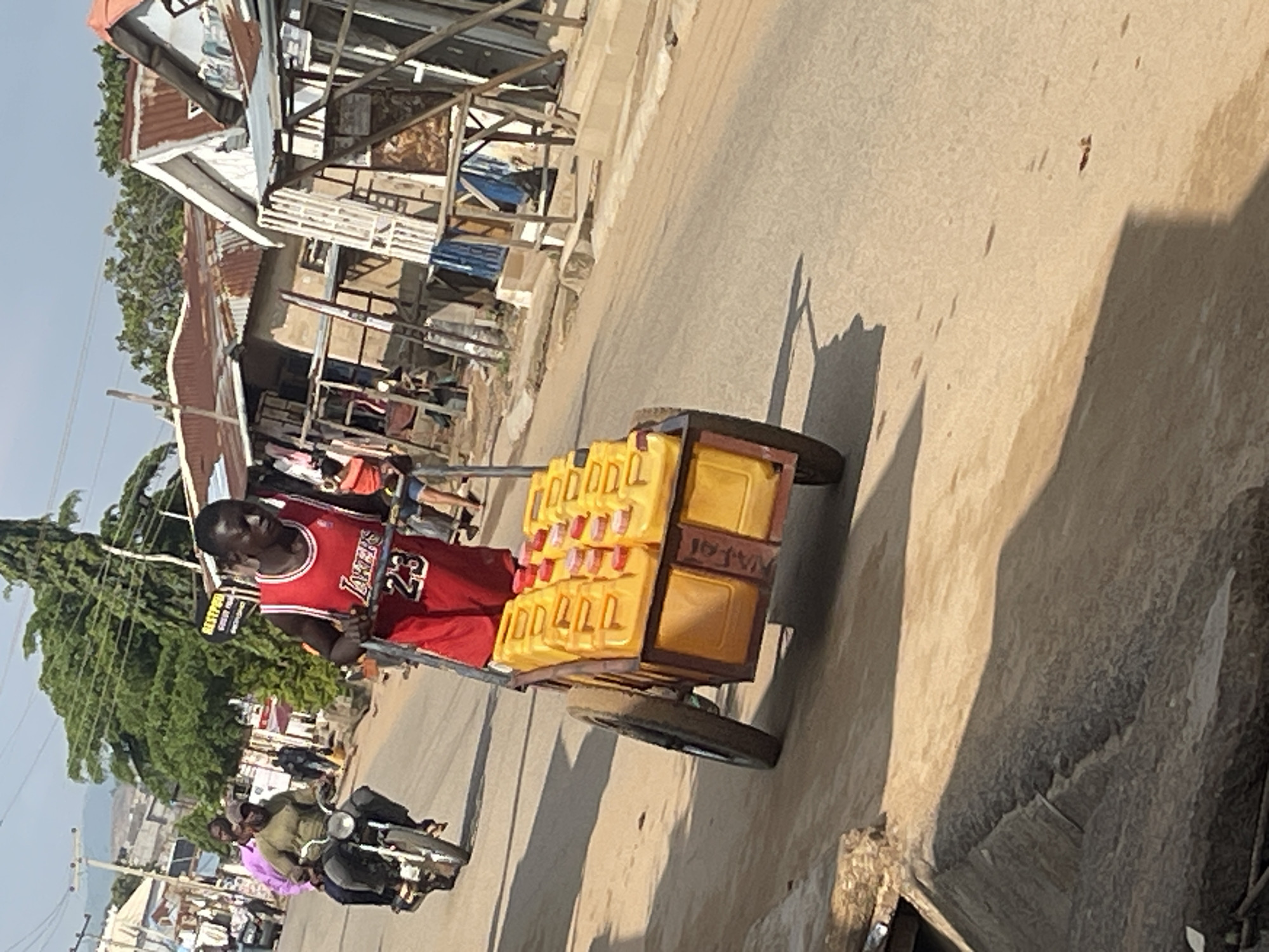 In a street scene in Abuja, Nigeria, a young man wearing a red basketball jersey with the number 23 is pushing a cart laden with yellow jerrycans filled with liquid. The cart has two large wheels and a metal frame. Behind him, two men are riding a motorcycle; the driver wears a green shirt and a cap, and the passenger wears a pink shirt. The background shows a street lined with buildings that appear to be market stalls or small shops, some with corrugated iron roofs. Trees and utility poles are also visible. The ground is a dusty, paved road. The lighting suggests it is daytime with bright sunlight, casting shadows on the road.