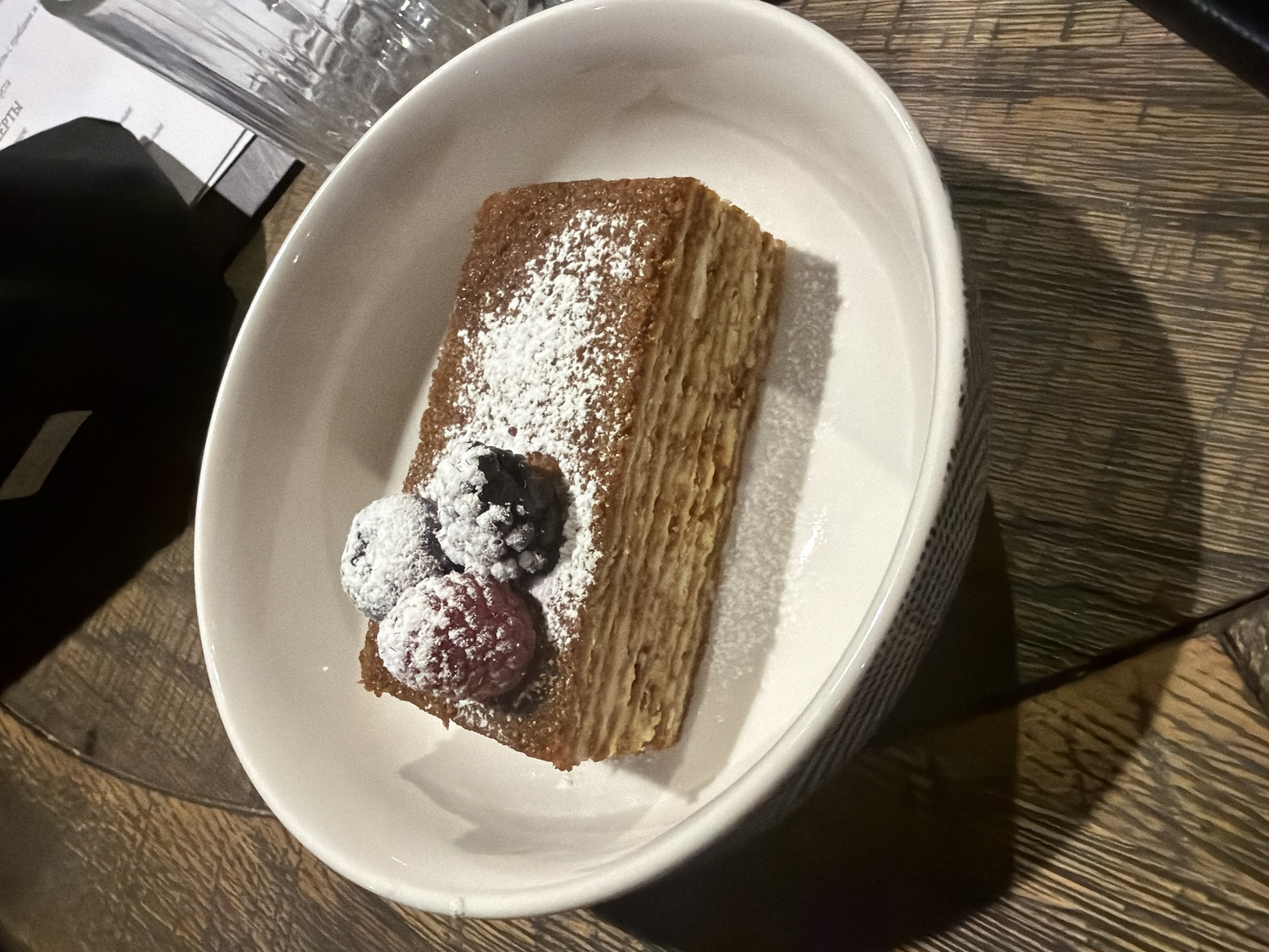 A dessert, likely a slice of layered cake dusted with powdered sugar and garnished with fresh berries, is presented in a white, oval-shaped dish with a patterned exterior. The dish is placed on a wooden table with a textured surface. To the left of the dish, out of focus, are parts of a beverage glass and a menu or card. The location is unspecified.
