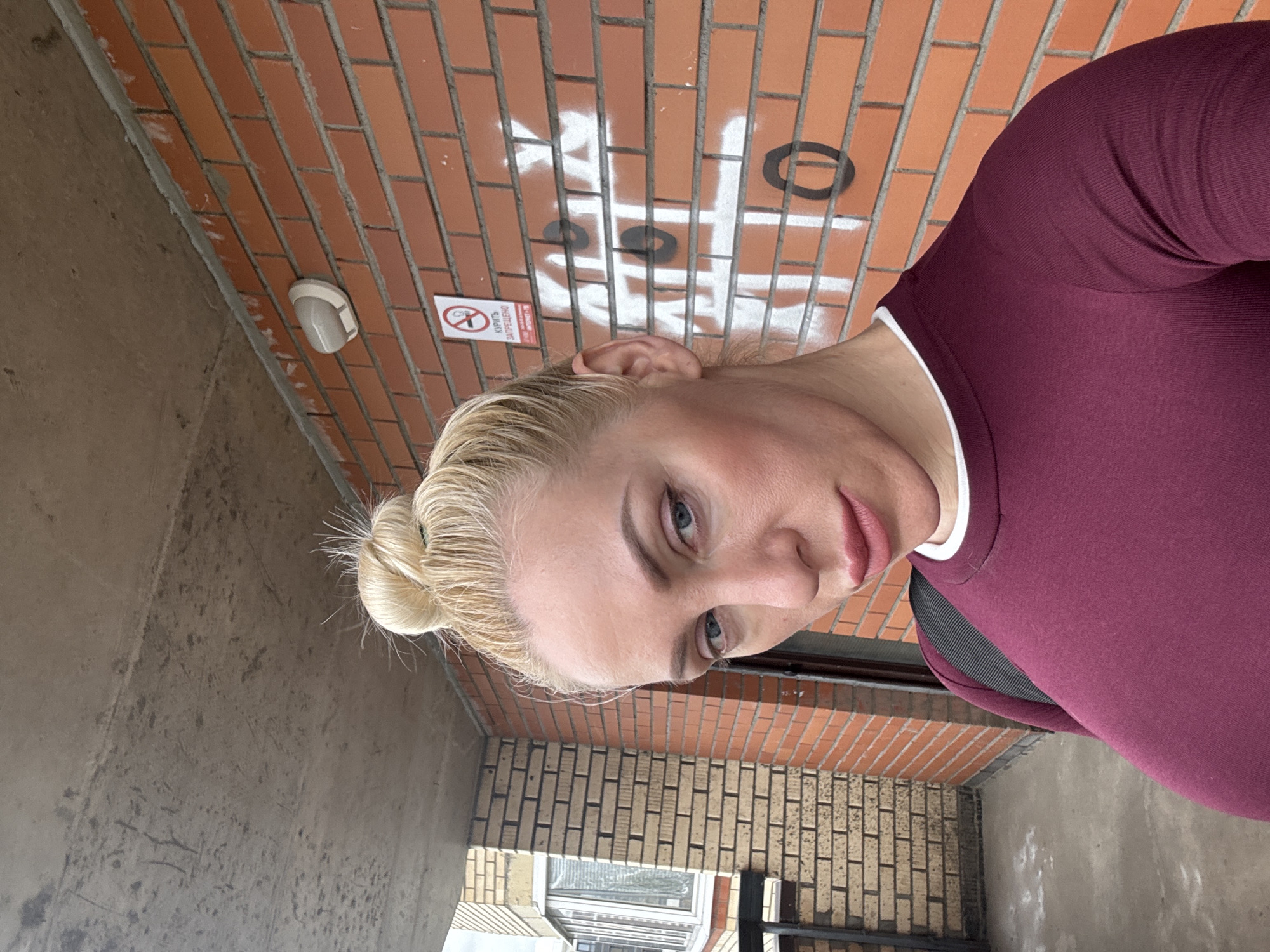 A woman with blonde hair in a bun is pictured outdoors, taking a selfie. She is wearing a maroon long-sleeved shirt with a white collar. Behind her is a brick wall with graffiti that appears to be a tic-tac-toe game. There is also a "No Smoking" sign on the wall. To the left of the woman is a building with a window. The ground is concrete.