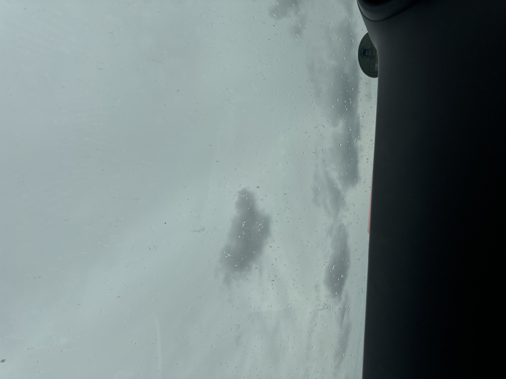 The image shows a view through a car windshield on a cloudy day. Raindrops are visible on the glass, distorting the view of a grey sky with scattered dark clouds. In the lower part of the image, the dark dashboard of a car is visible. There is also a glimpse of what appears to be a sticker or decal on the upper right portion of the dashboard. The scene depicts a typical overcast day, possibly during or after rainfall, consistent with the weather often experienced in Tinryland, Ireland. There are no people or specific landmarks visible that would pinpoint a precise location within the city.