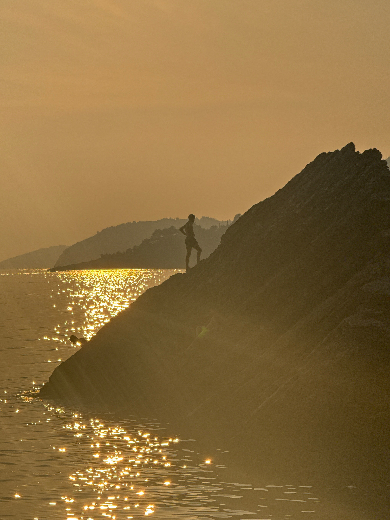 The image depicts a silhouetted person standing on a rocky outcrop overlooking a body of water. The sun is low in the sky, casting a warm, golden glow that creates shimmering reflections on the water's surface. In the background, hazy hills rise from the water, also silhouetted against the sky. The scene appears to be the coast of Kamenovo, Montenegro, with the rocky terrain and the reflective water characteristic of the Adriatic Sea. The overall impression is one of tranquility and natural beauty, possibly captured during sunset.