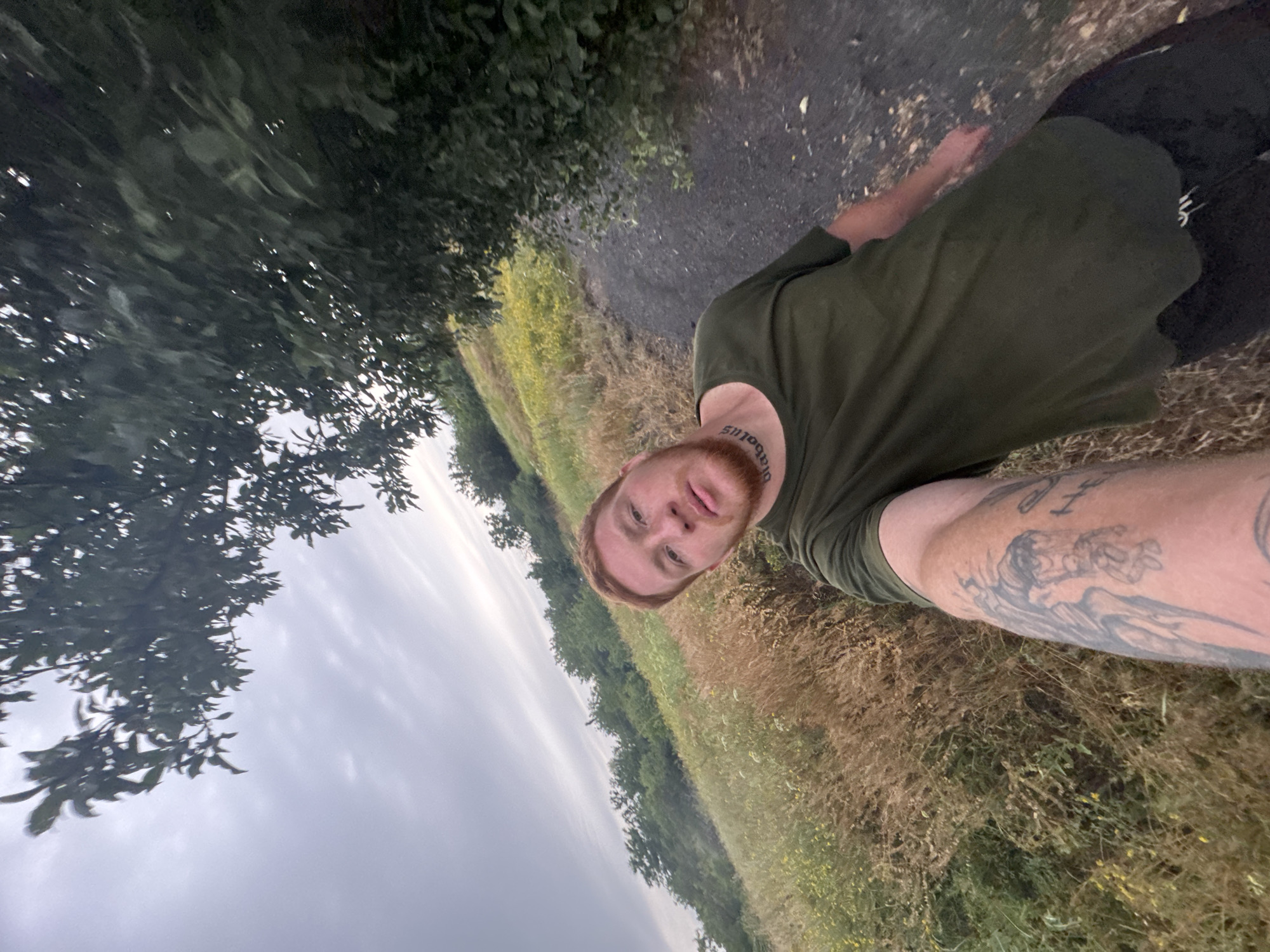 In a low-angle selfie taken outdoors, a man with ginger hair and a beard faces the camera. He wears a dark green t-shirt and black pants. A tattoo is visible on his left arm, and another tattoo reads "diabolus" on his neck. The man is standing on a dark path next to a field of dry grass and some yellow wildflowers. Trees and foliage surround the path and the field. The sky is overcast and gray. The location is listed as Unknown, Unknown.