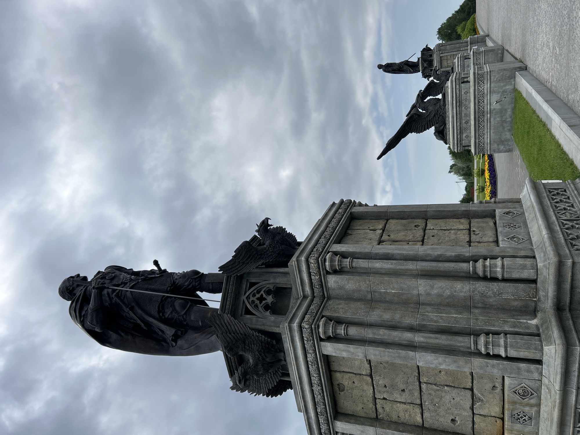 The image shows a series of statues in Khryashchevka, Russia. The primary subject is a large, dark statue of a man in armor and a cape, holding a sword. This statue stands on a multi-tiered stone pedestal adorned with architectural details and two griffin-like creatures. In the background, receding into the distance, are other similar statues, each featuring a griffin and a figure, spaced along a paved walkway and grassy area. The sky is overcast with dramatic clouds. The statues appear to be part of a historical or mythological monument.