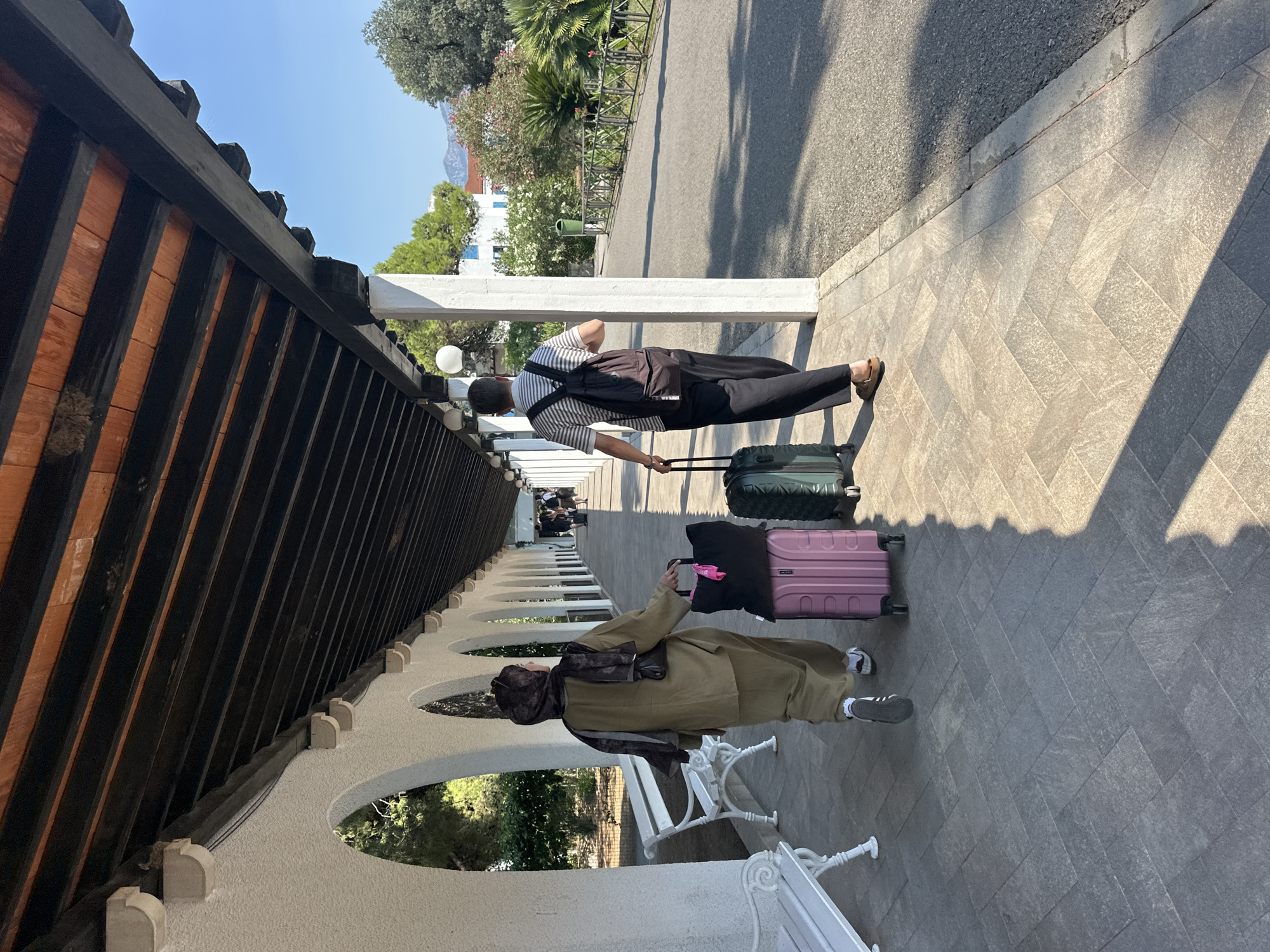 Two individuals are walking away from the camera on a paved walkway lined with arched white structures and benches, beneath a dark wooden overhang. The person on the left wears a hijab and an olive-green matching outfit, pulling a pink suitcase and carrying a black bag. The person on the right wears a striped t-shirt, dark pants, and a backpack, pulling a green suitcase. The walkway leads to an outdoor area with a road, greenery, and buildings in the background, under a clear blue sky. This appears to be the outdoor walkway of a hotel or resort in Budva, Montenegro.