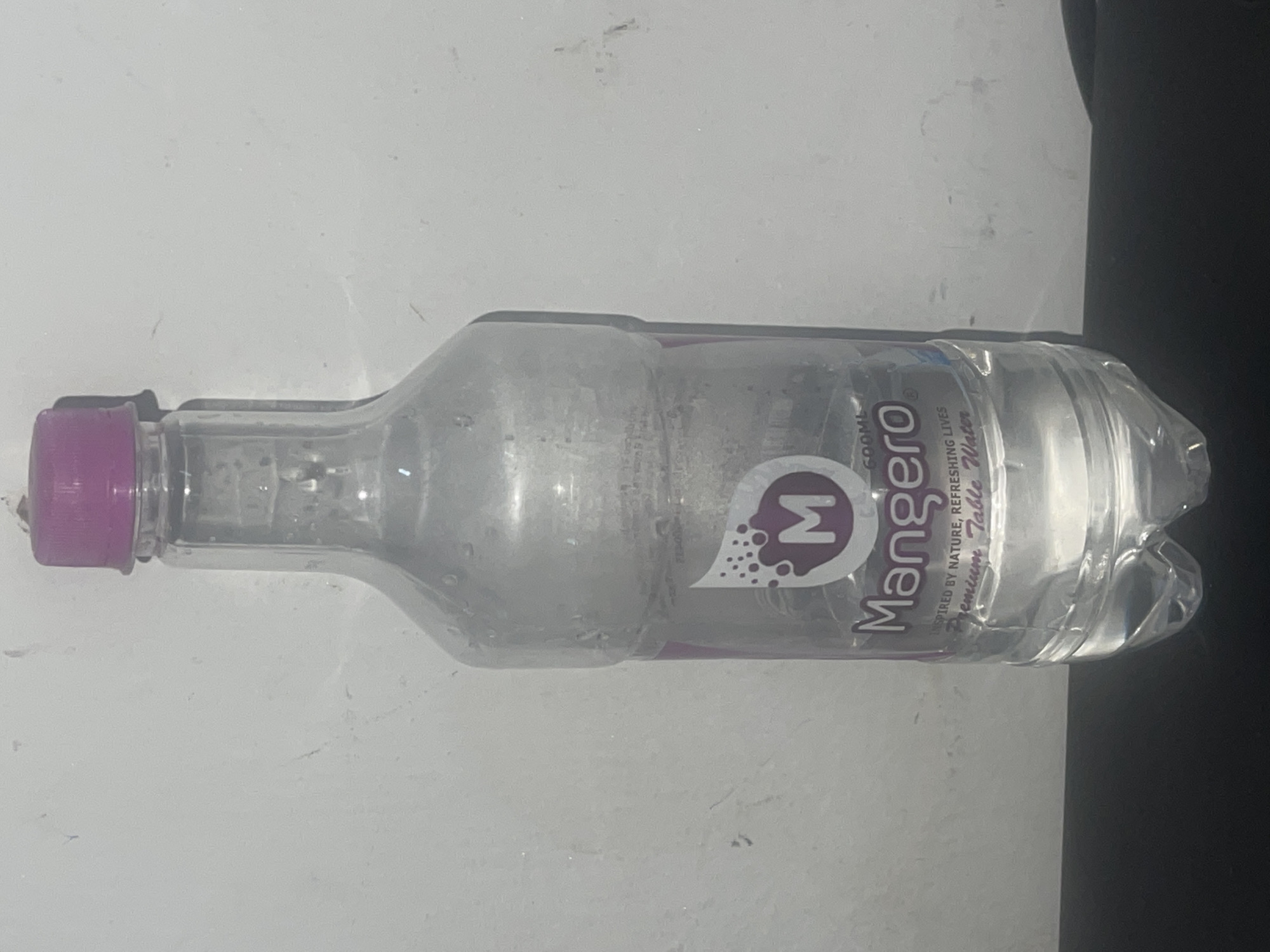 A half-empty, clear plastic bottle of "Mangero" premium table water with a purple cap and label is lying horizontally on a white surface. The label features a stylized letter "M" and the word "Mangero". The background is plain and light. The image was taken in Abraka, Nigeria.