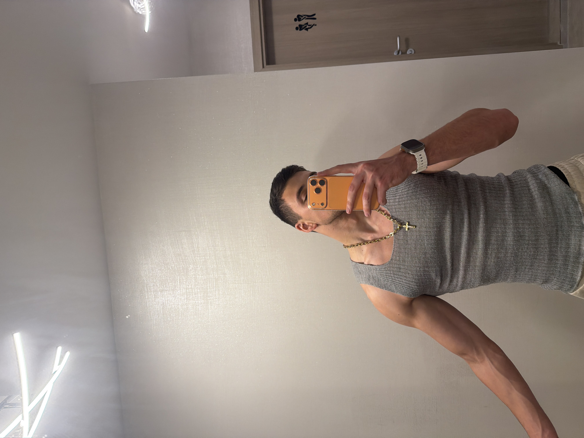 The original image is rotated 90 degrees counter-clockwise. When viewed upright, it depicts a man taking a mirror selfie in an indoor setting. The man, with short dark hair and a neat beard, is positioned centrally, seen from the chest up. He holds an orange-cased smartphone to his face, partially obscuring his eyes and nose. He is wearing a grey ribbed tank top, a thick gold chain with a prominent cross pendant, and a white-banded smartwatch on his left wrist. His right arm is visible, showing prominent veins. The bottom portion of light-colored shorts or pants can be seen.
The background is a reflection in the mirror, revealing a room with light-colored, possibly textured walls that show a subtle sparkle. In the upper right of the reflection, there is a light-colored wooden door with black stylized figures representing restrooms (one seated, one standing). Bright artificial light sources are present in the reflection; one is a visible light fixture at the top center, and another, more prominent, elongated light source is on the lower left side. The image captures the moment of a casual selfie. No visible text is present beyond the stylized restroom symbols.