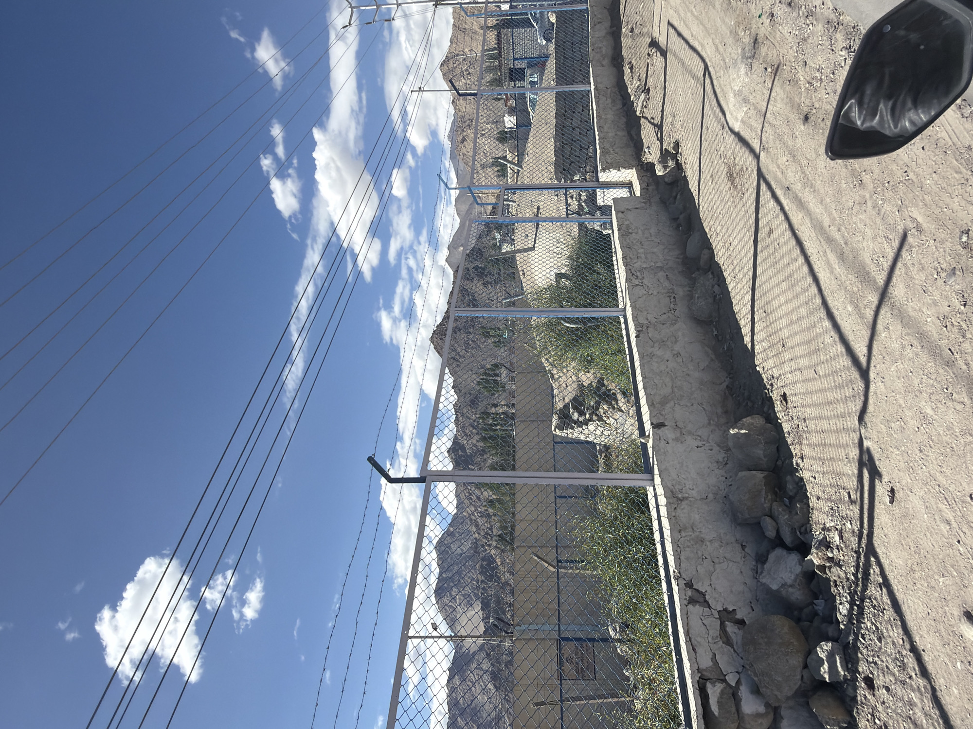 The image depicts a view of a street in Leh, India, with a chain-link fence in the foreground and a mountainous landscape in the background. The fence is topped with barbed wire and is supported by metal posts. Beyond the fence, there are some structures that appear to be buildings, possibly part of a settlement. The ground is unpaved and dusty, with some rocks and debris scattered around. Power lines are visible crossing the sky, which is blue with scattered white clouds. The overall impression is of an arid, high-altitude environment.