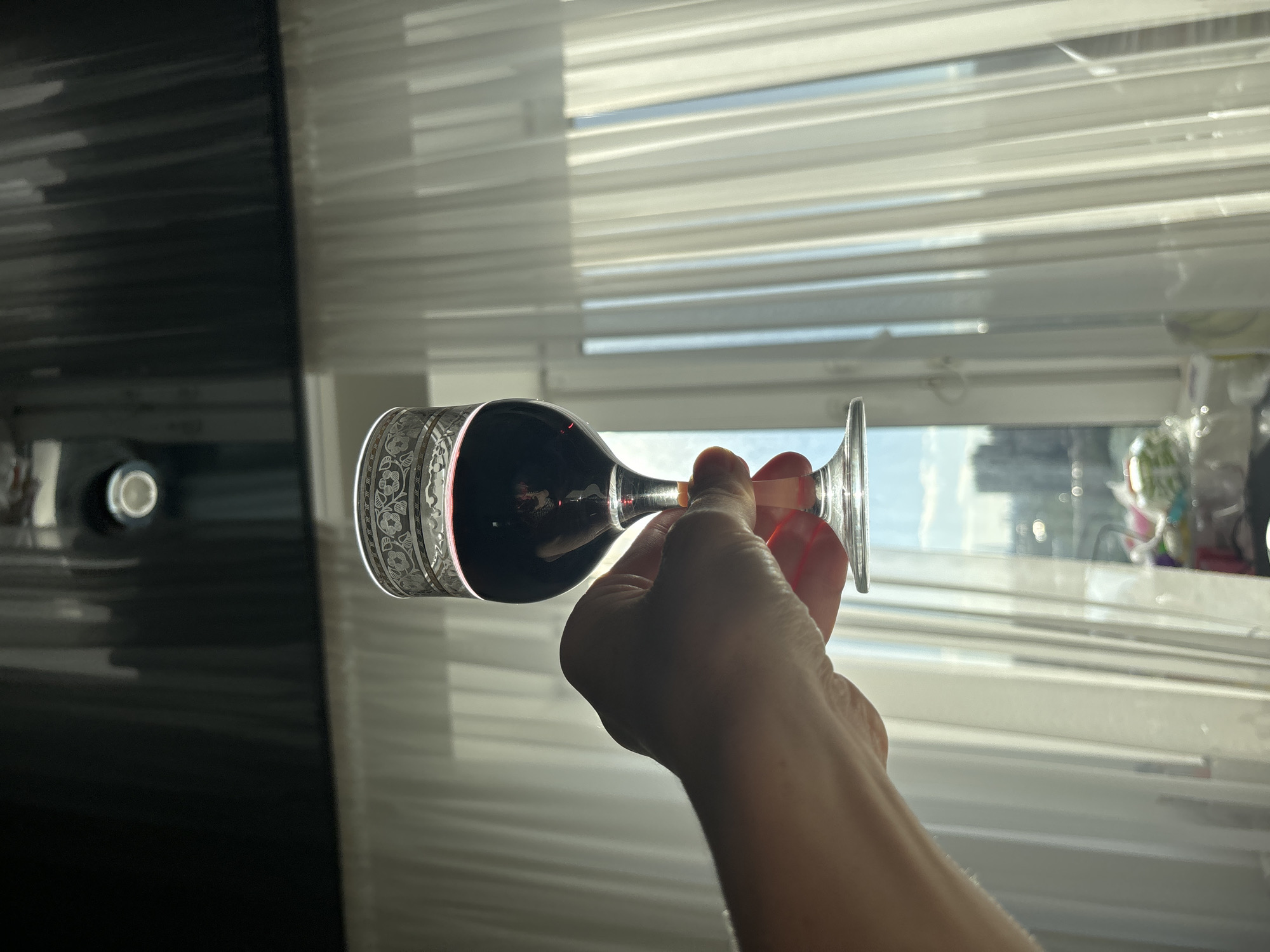 A hand holds a glass of red wine against a backdrop of sheer white curtains, allowing sunlight to stream through. The glass has a decorative silver band around its rim. Beyond the curtains, a blurry urban landscape with buildings and trees is visible. The ceiling above is dark and reflective, with a circular light fixture. The scene appears to be indoors, likely in a residential setting.