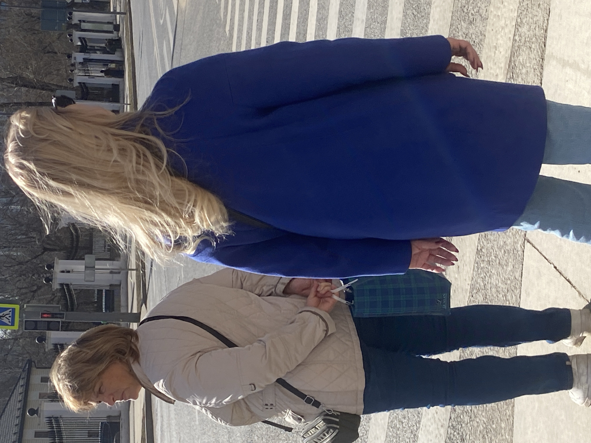 Two women stand on a paved surface in a city environment. The woman closest to the viewer wears a light-colored quilted jacket, dark pants, and white sneakers. She has short, light brown hair. A cross-body bag with a silver and black design hangs at her side. The woman behind her is taller, wearing a bright blue coat and blue jeans. She has long blonde hair. They are standing on a gray surface with light gray stripes, likely a crosswalk. A traffic light and pedestrian crossing sign are visible in the background. The scene appears to be Moscow, Russia, suggested by the street furniture and overall urban atmosphere.
