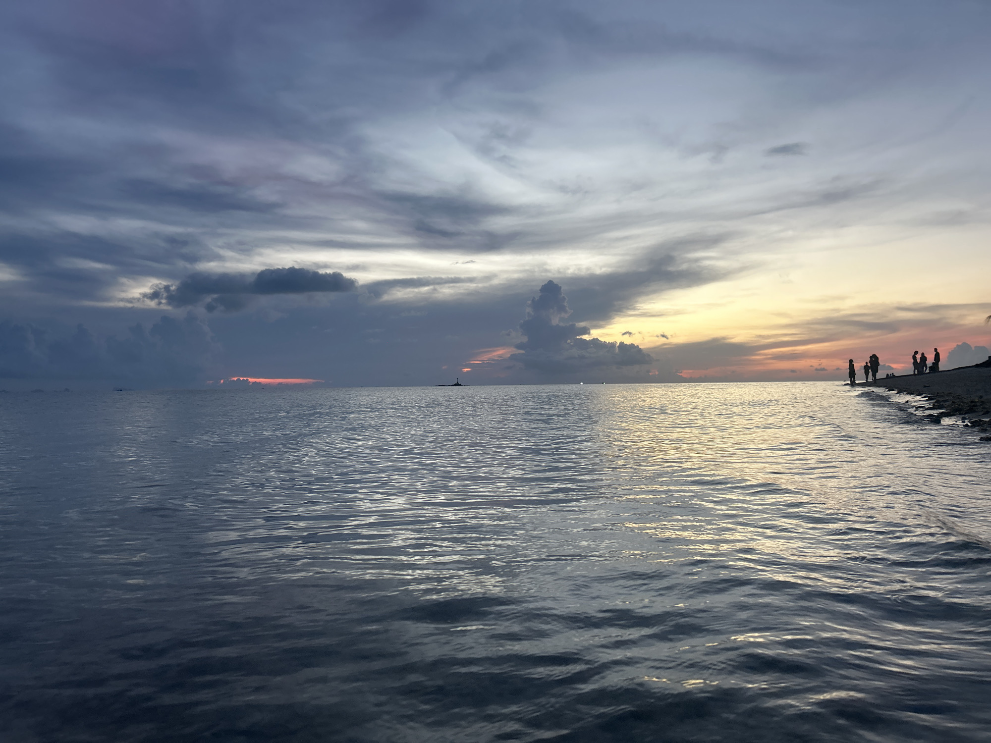 The image depicts a seascape at sunset in Baan Sri Thanu, Thailand. A group of silhouetted people stand on the beach to the right. The ocean occupies most of the scene, with gentle waves reflecting the light of the setting sun. In the distance, a small structure, possibly a lighthouse or building, is visible on the horizon. The sky is filled with clouds, ranging from dark grey to lighter shades with orange hues where the sun is setting.