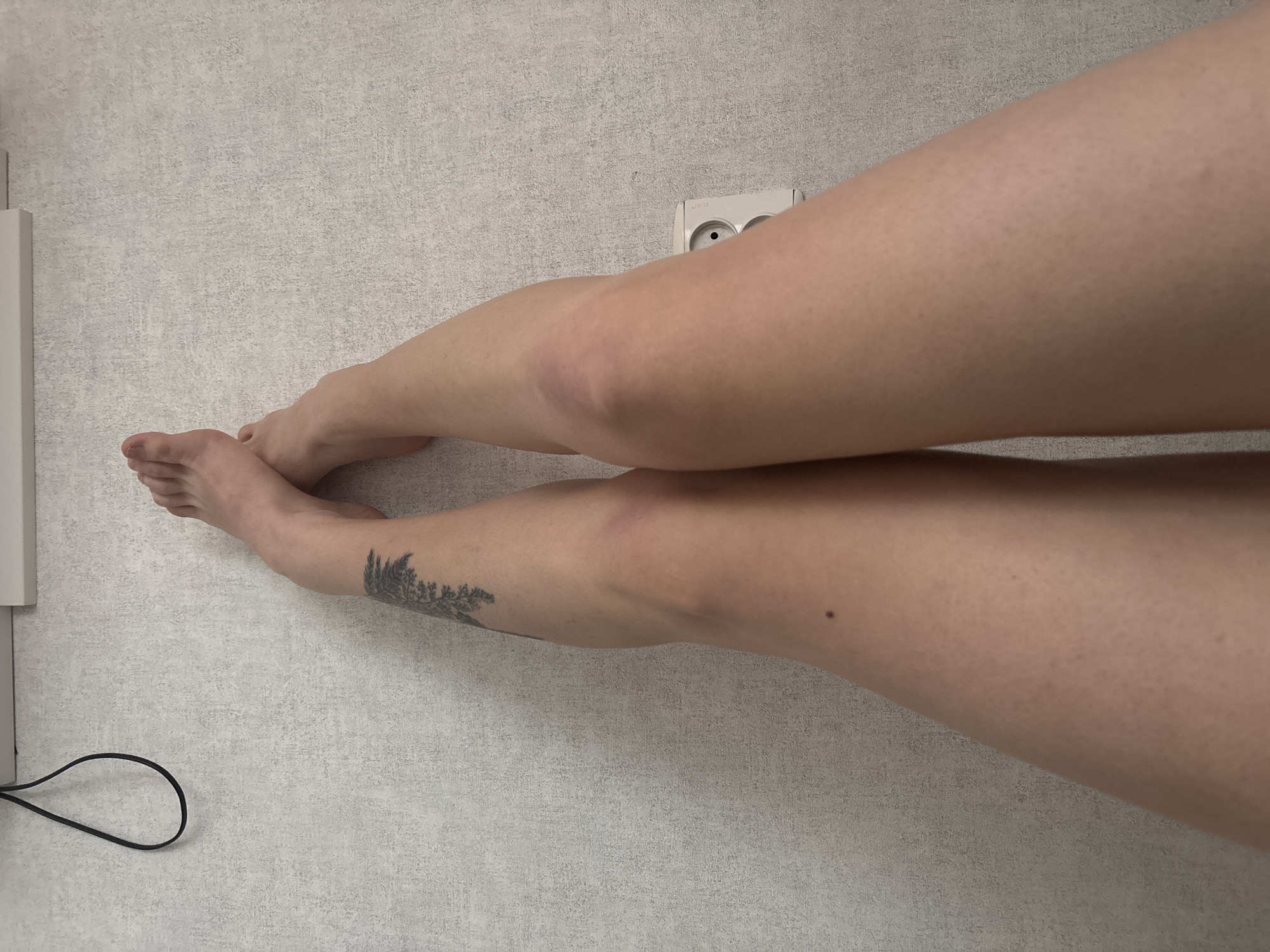 The image shows a person's legs crossed at the ankles, resting against a textured, light-colored wall. The person has fair skin and a tattoo of a fern or tree branch on their left calf. A black cable hangs loosely in the upper left corner of the frame. To the right of the legs, a white electrical outlet with two sockets is visible on the wall. The lighting is soft and diffused, suggesting an indoor environment. The context of Krasnoyarsk, Russia is not visually evident from the image itself.