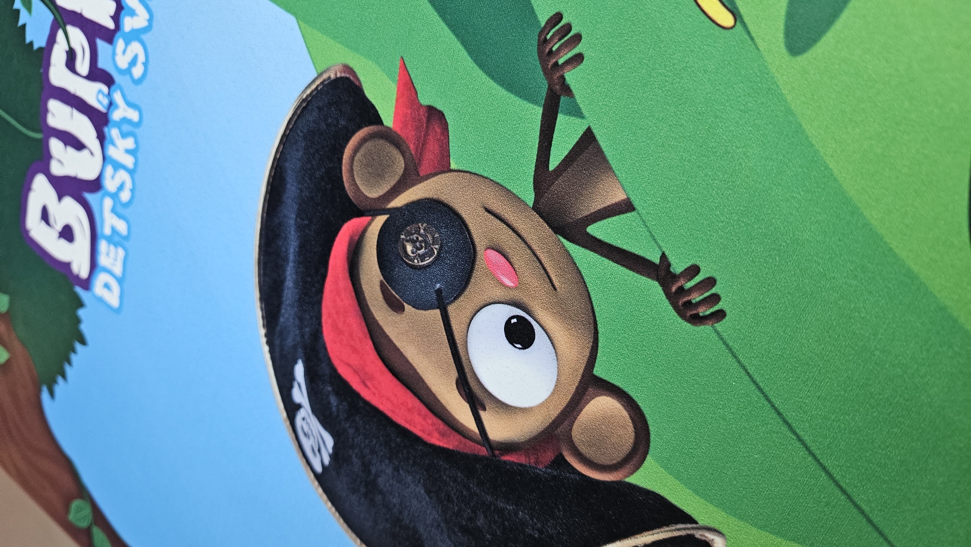 The image shows a close-up of a cartoon monkey dressed as a pirate. The monkey has brown fur, a black pirate hat with a skull and crossbones, a red bandana, and a black eyepatch over its right eye. It has large white eyes with black pupils, a small pink nose, and is smiling. Its arms and hands are visible, reaching out from behind a bright green surface, which appears to be a leaf or a hill. In the upper part of the image, there is text that reads "BURFI DETSKY SV". The background is a bright blue sky with a brown tree branch and green leaves at the top left. This appears to be a detail from a poster or advertisement, likely for a children's show or product, possibly related to Bratislava, Slovakia given the text.